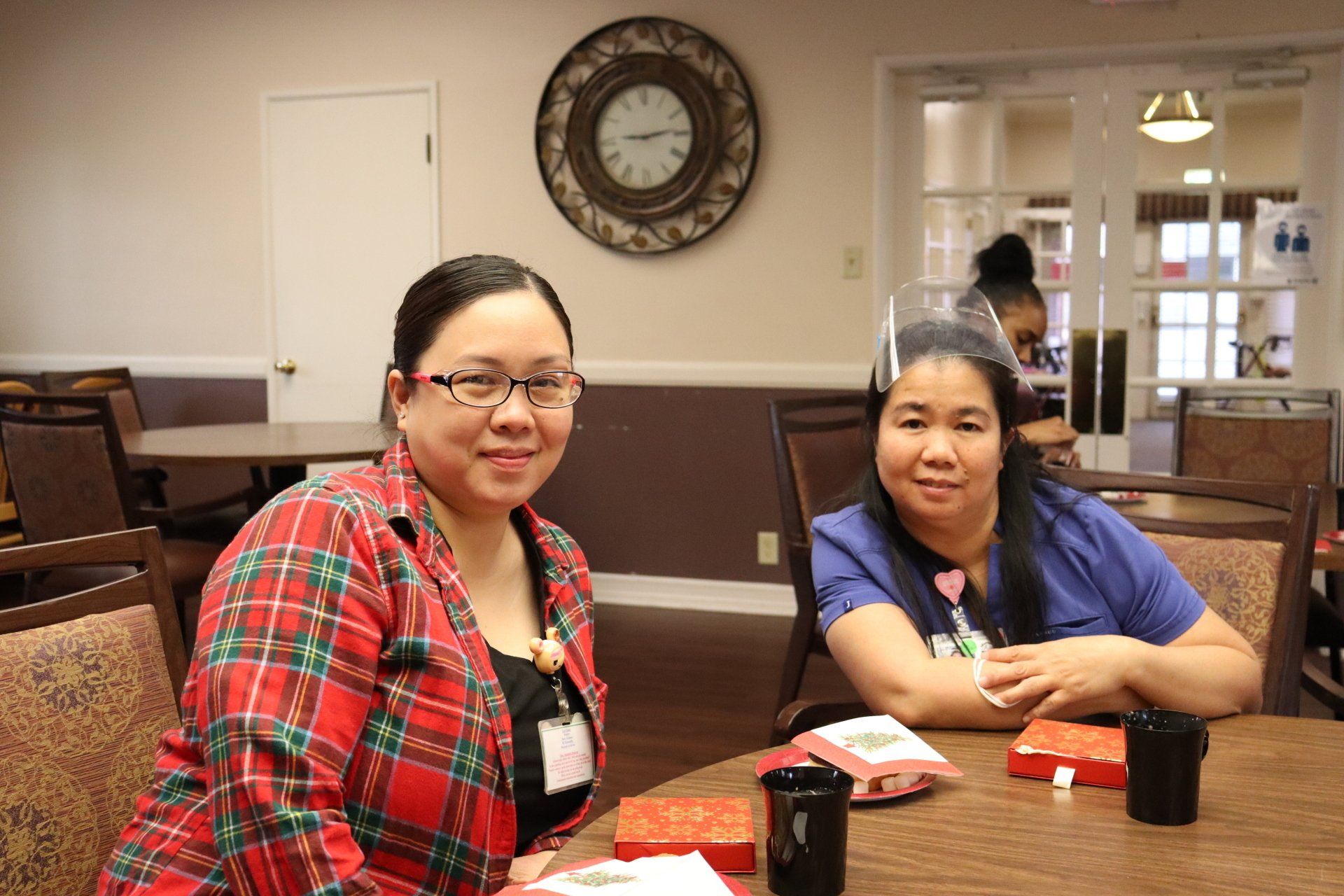 Two women sitting at a table, smiling. Indoors with a clock and door visible. One wears a red plaid shirt.