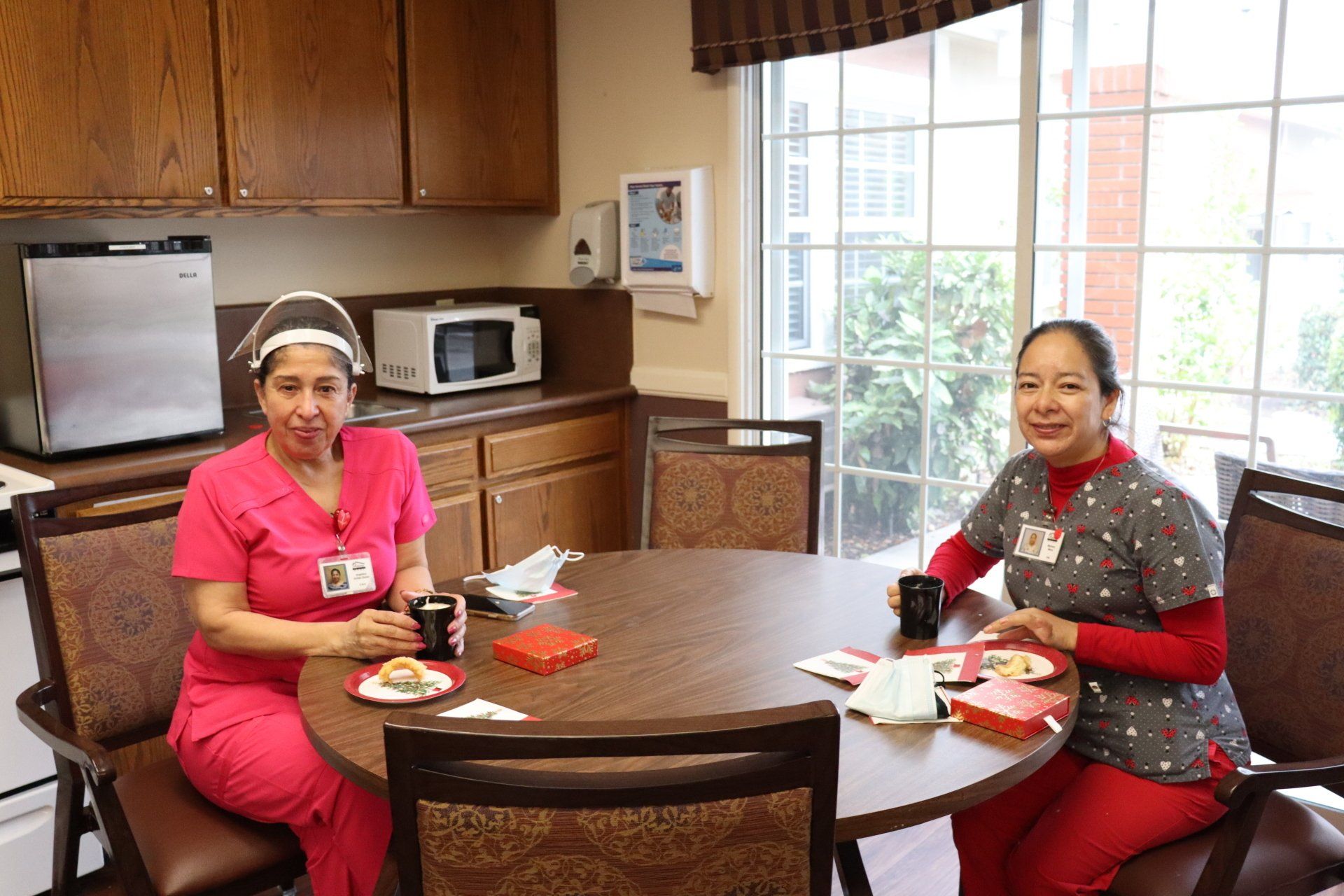 Two women in scrubs sit at a round table in a kitchen, drinking coffee.