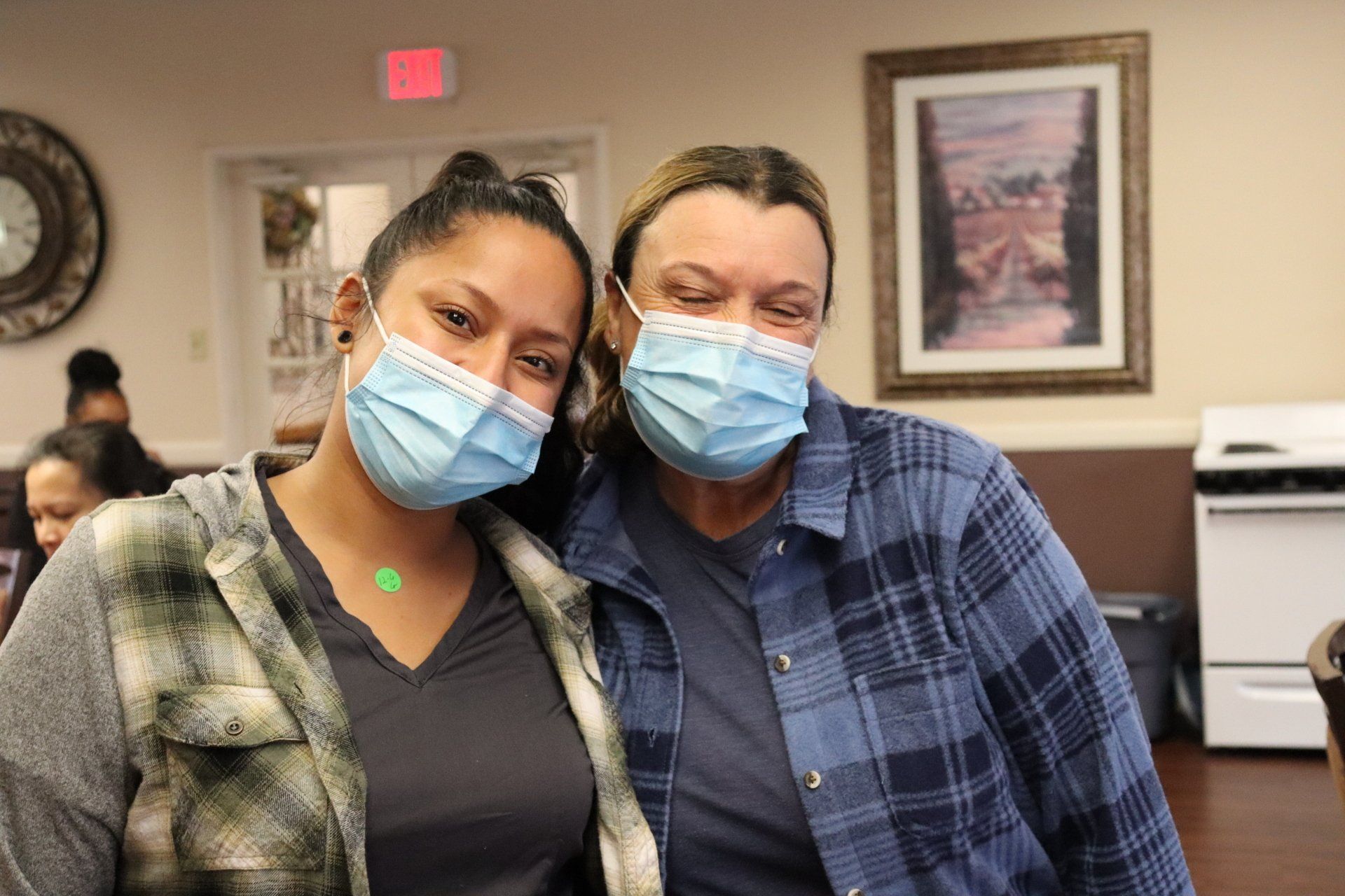 Two women wearing masks pose indoors; one in plaid, the other with a green-gray jacket.