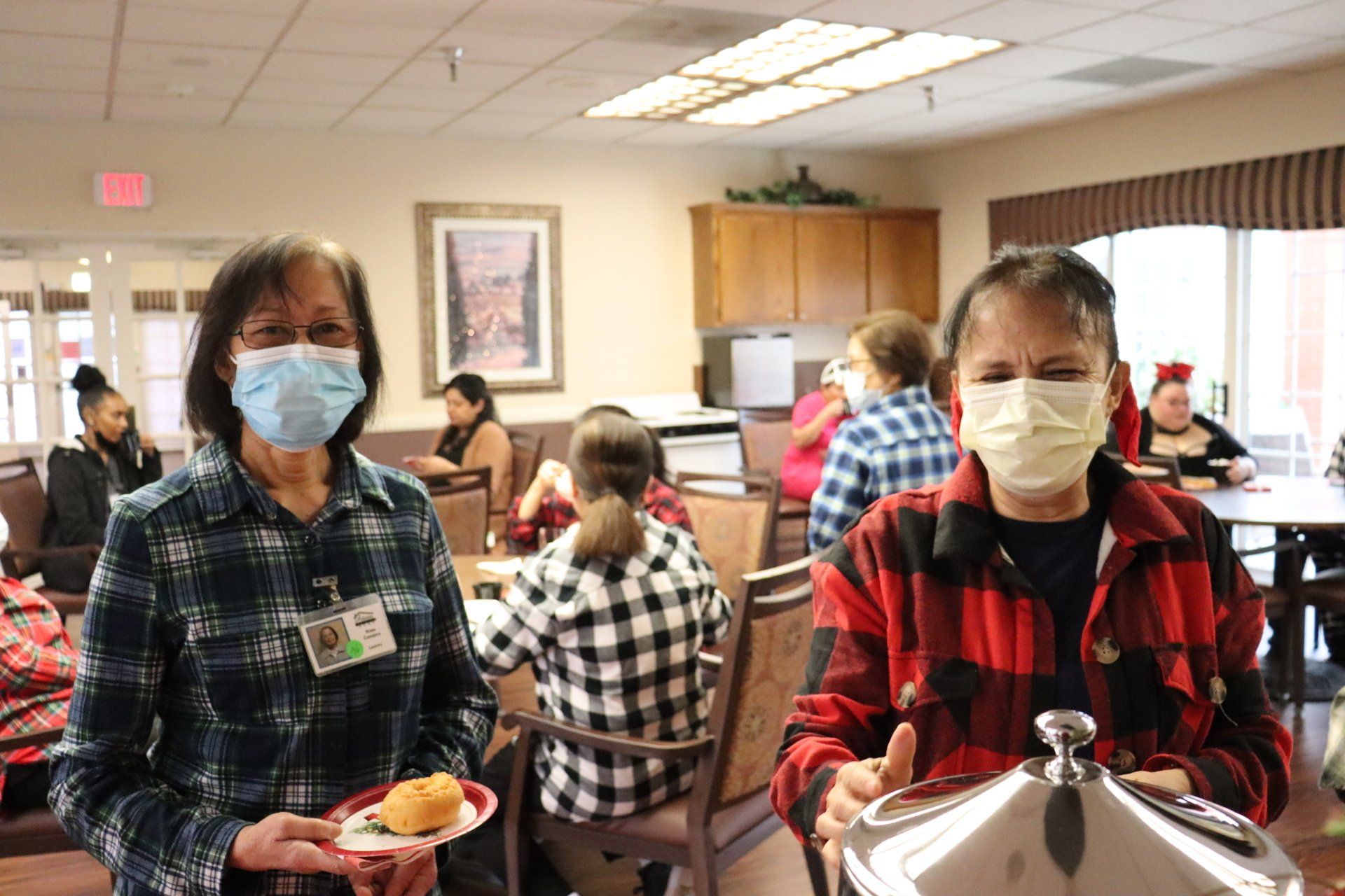 Two masked women with food trays in a dining hall; others seated at tables.