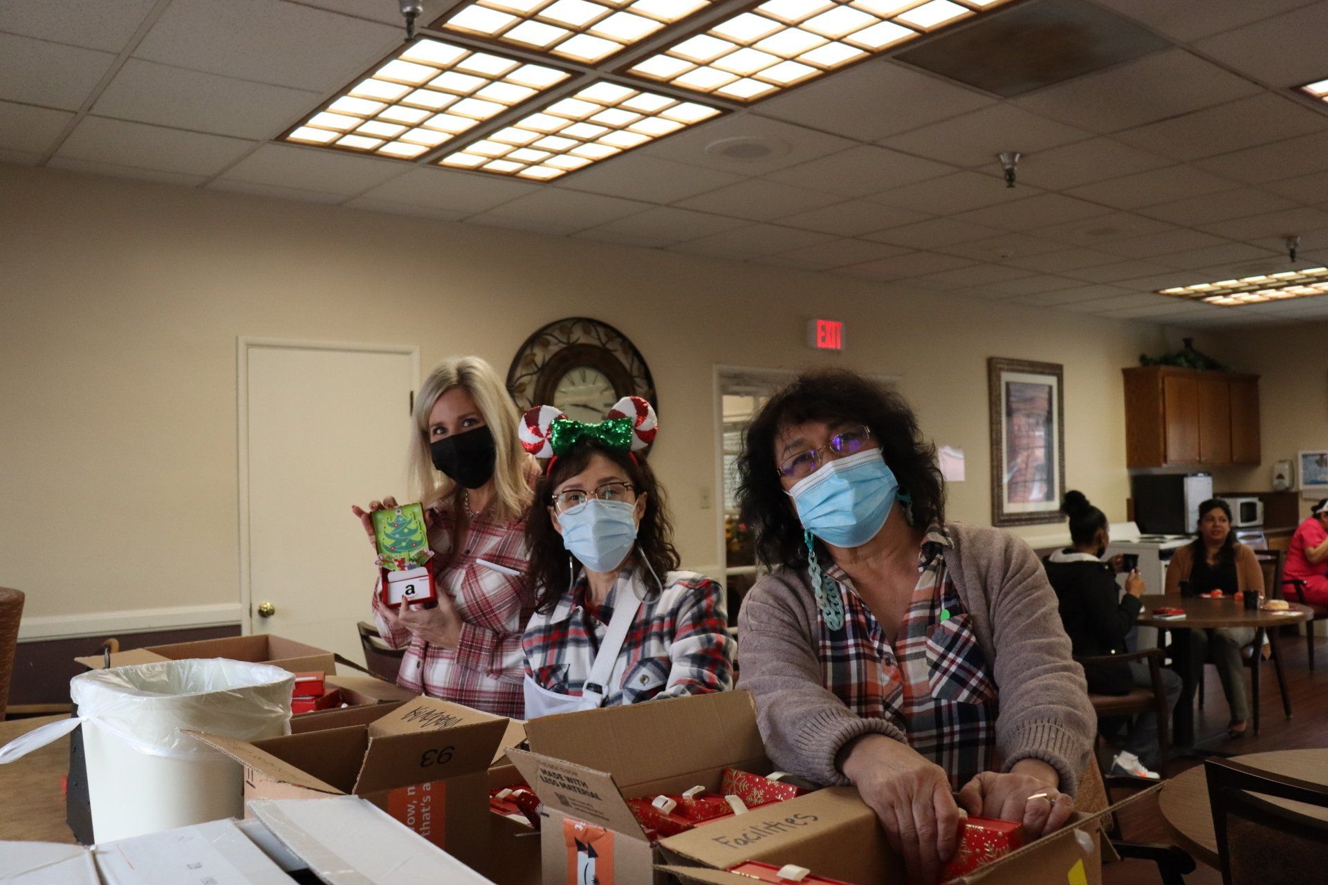 Three women wearing masks pack boxes, holiday themed, indoors.