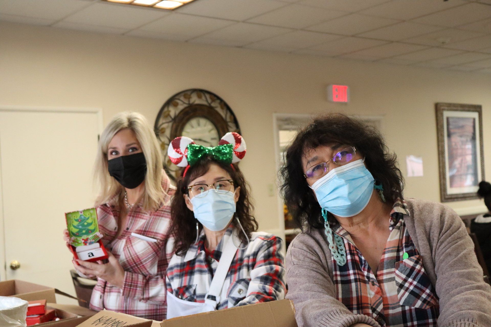 Three women wearing masks and plaid shirts preparing gifts in a room.