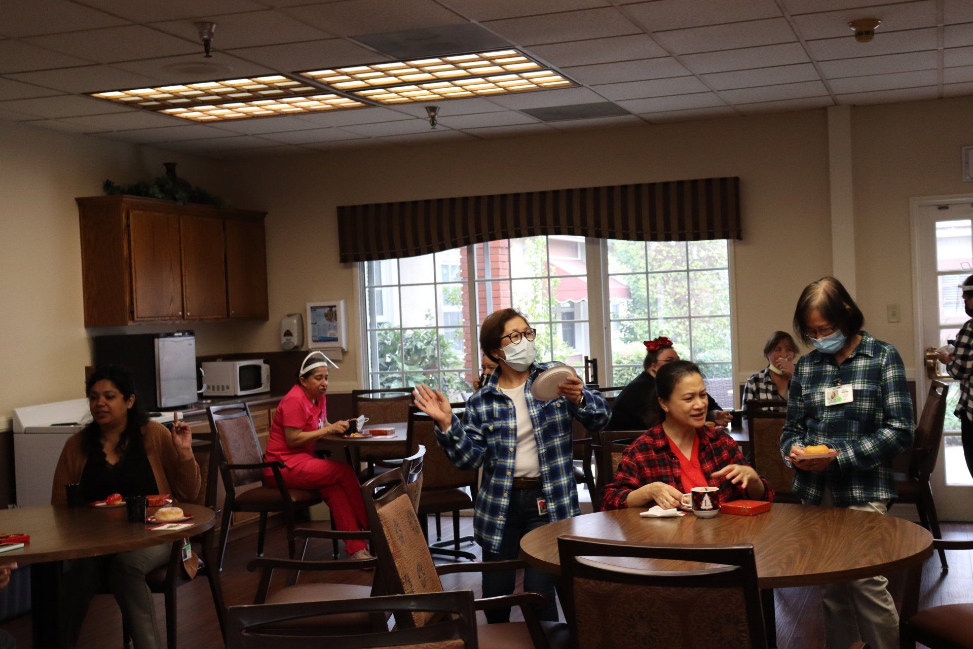 People in a dining room, some wearing masks. One woman gestures excitedly. Others sit at tables, some playing games.