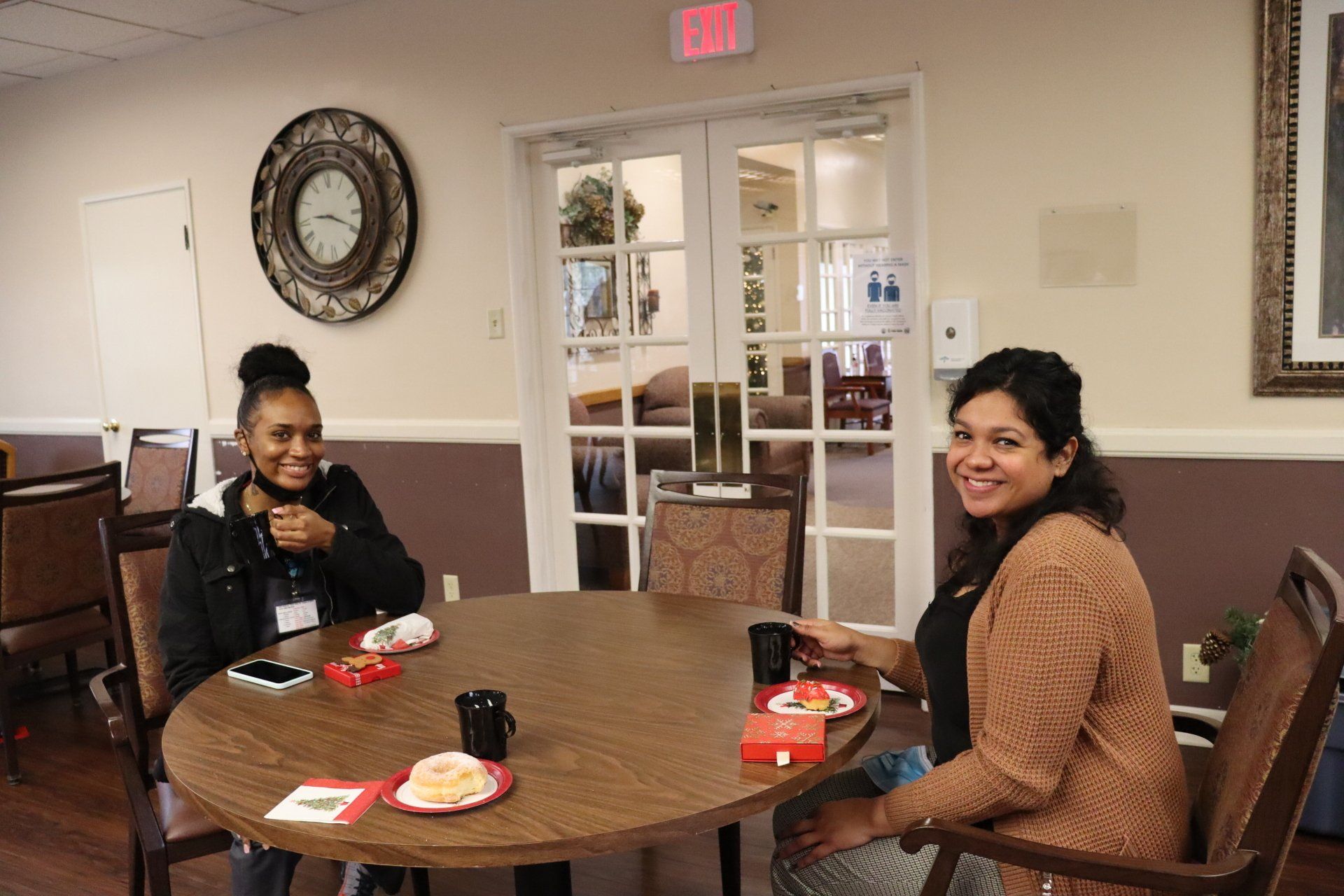 Two women smiling, sitting at a round table. One has black hair, the other is brown-haired, both holding mugs indoors.