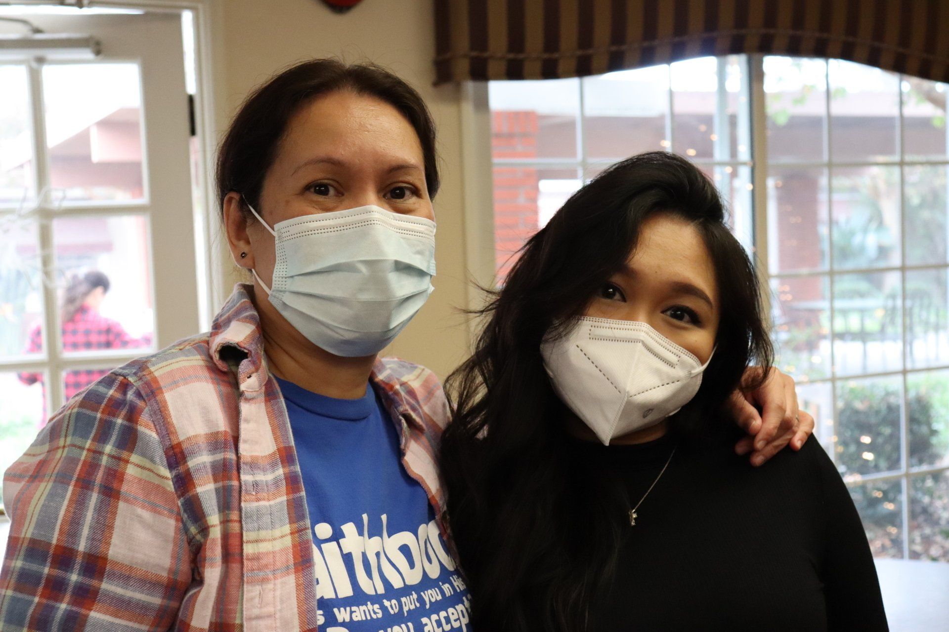 Two masked women smiling, arm around each other, near a window.