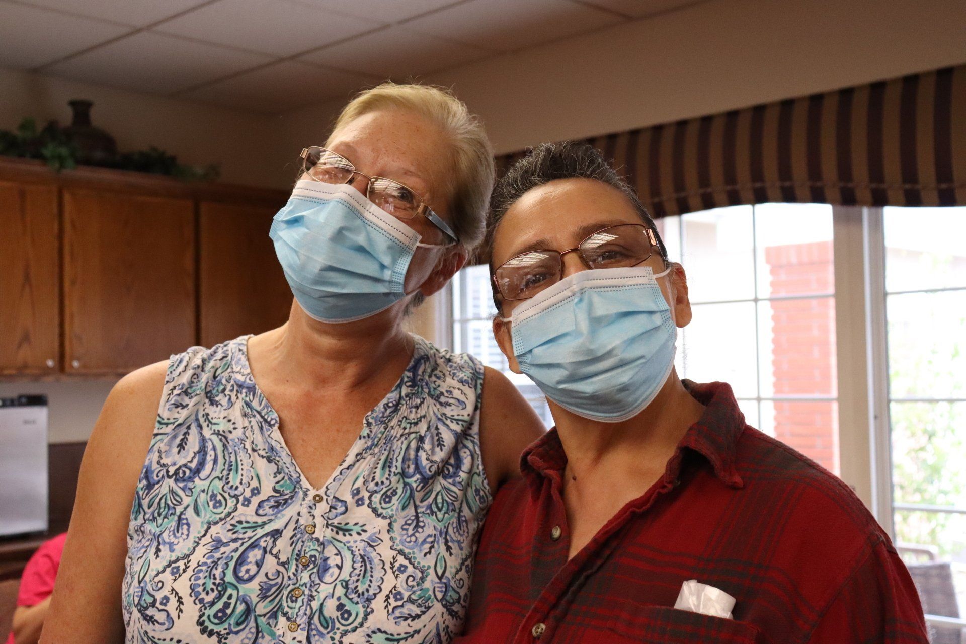 Two women in masks pose together, looking at the camera. One wears a patterned top, the other a red plaid shirt.