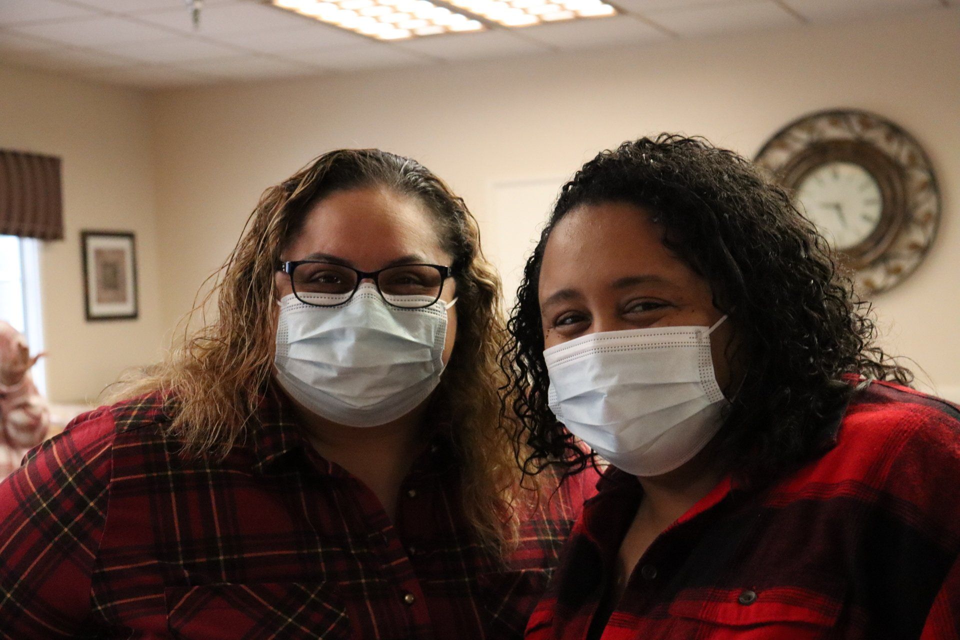 Two women wearing masks, posing indoors, one with glasses, both in red plaid shirts.