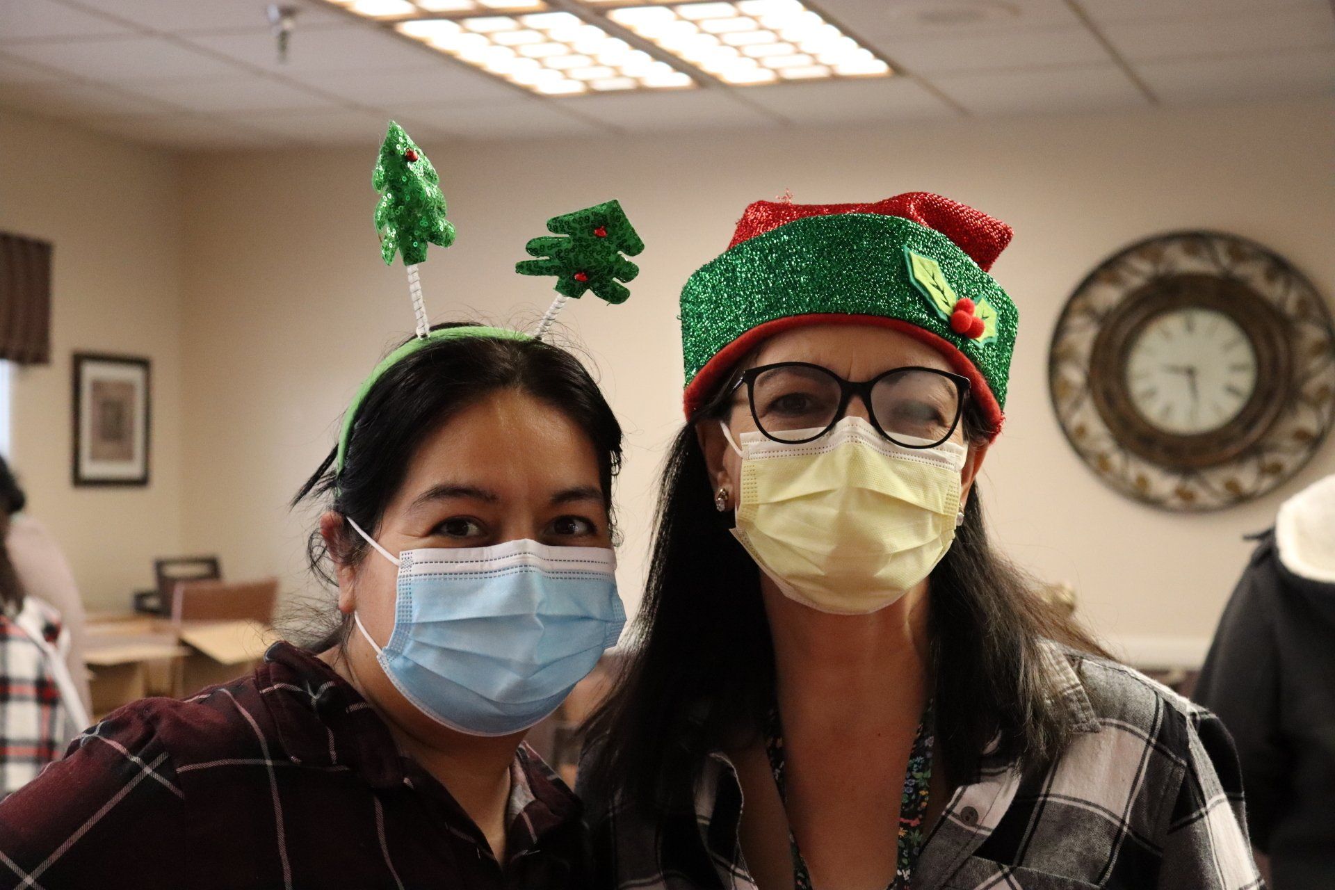 Two women wearing masks and Christmas-themed headgear smile indoors.