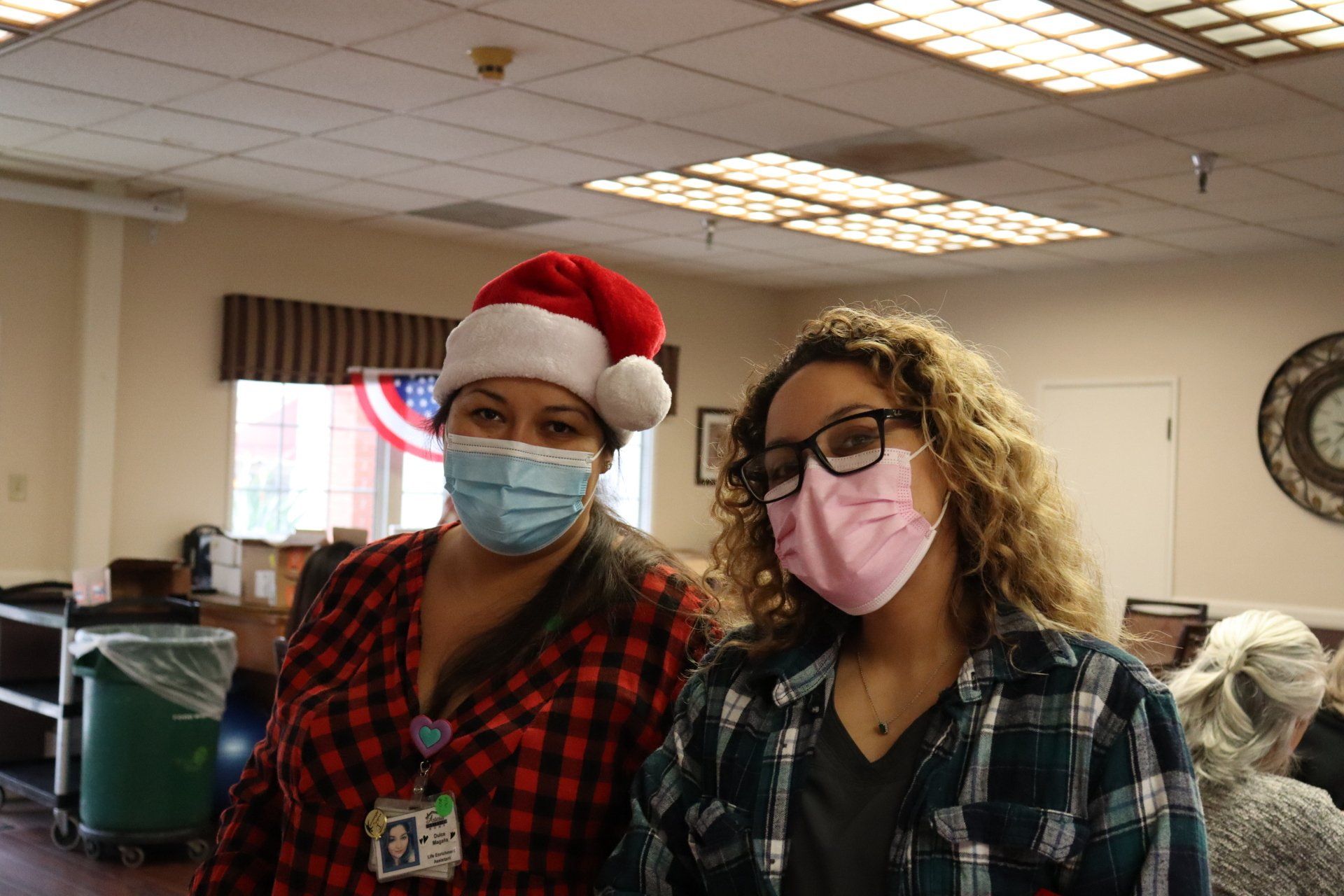 Two women in masks, one with a Santa hat. They are smiling indoors, likely at a holiday event.