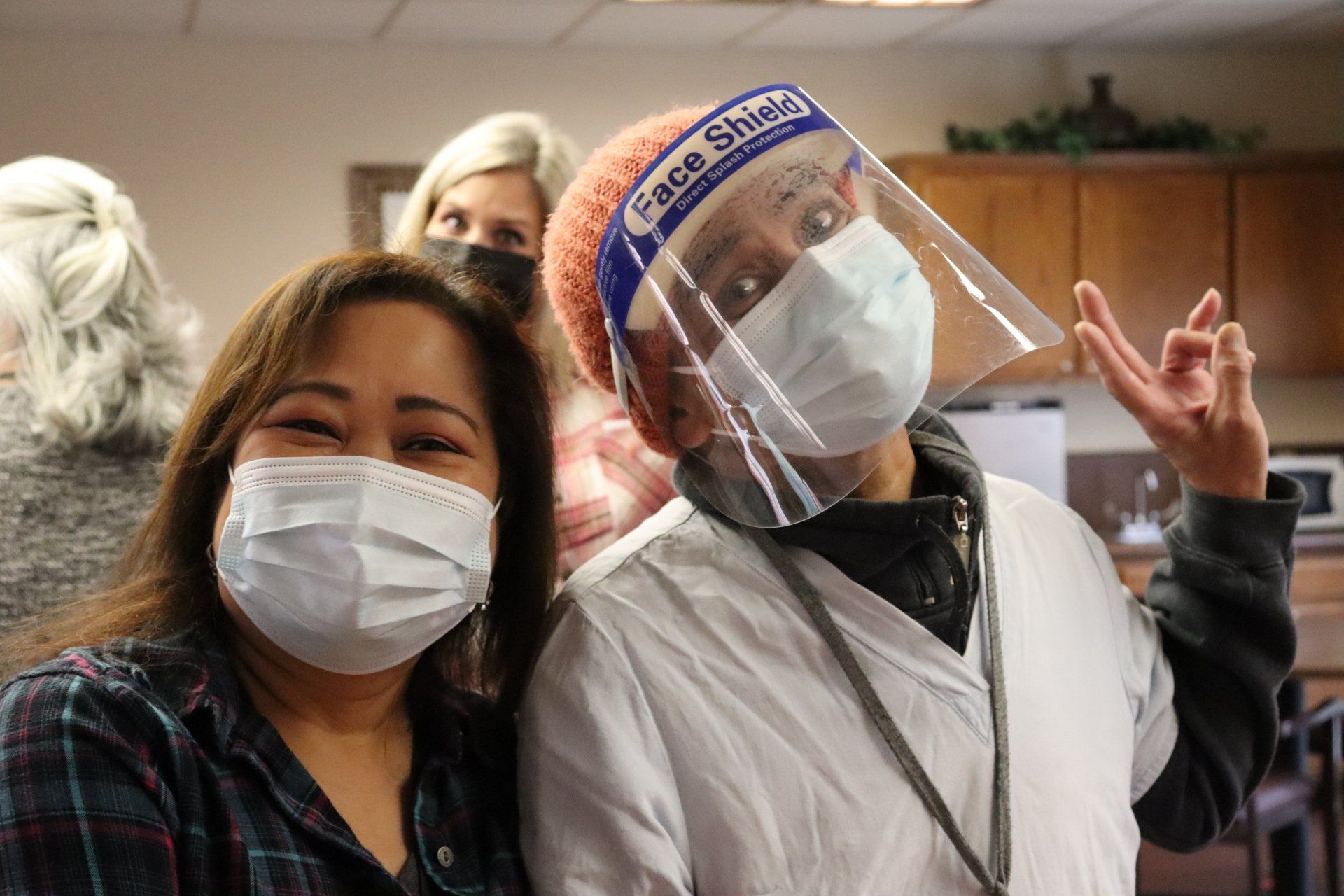 Two masked women smile. One wears a face shield. Others in the background. Indoor setting.