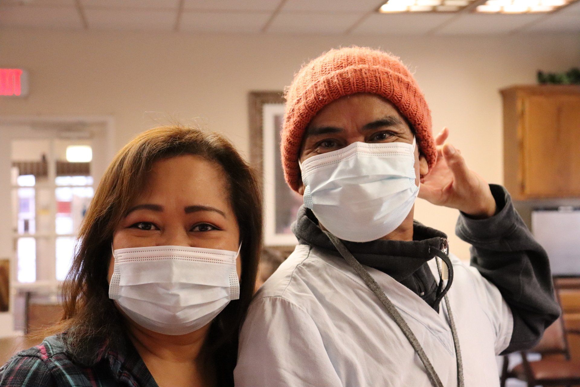 Two people wearing face masks indoors. Woman on left, man on right adjusting his mask; both smiling.