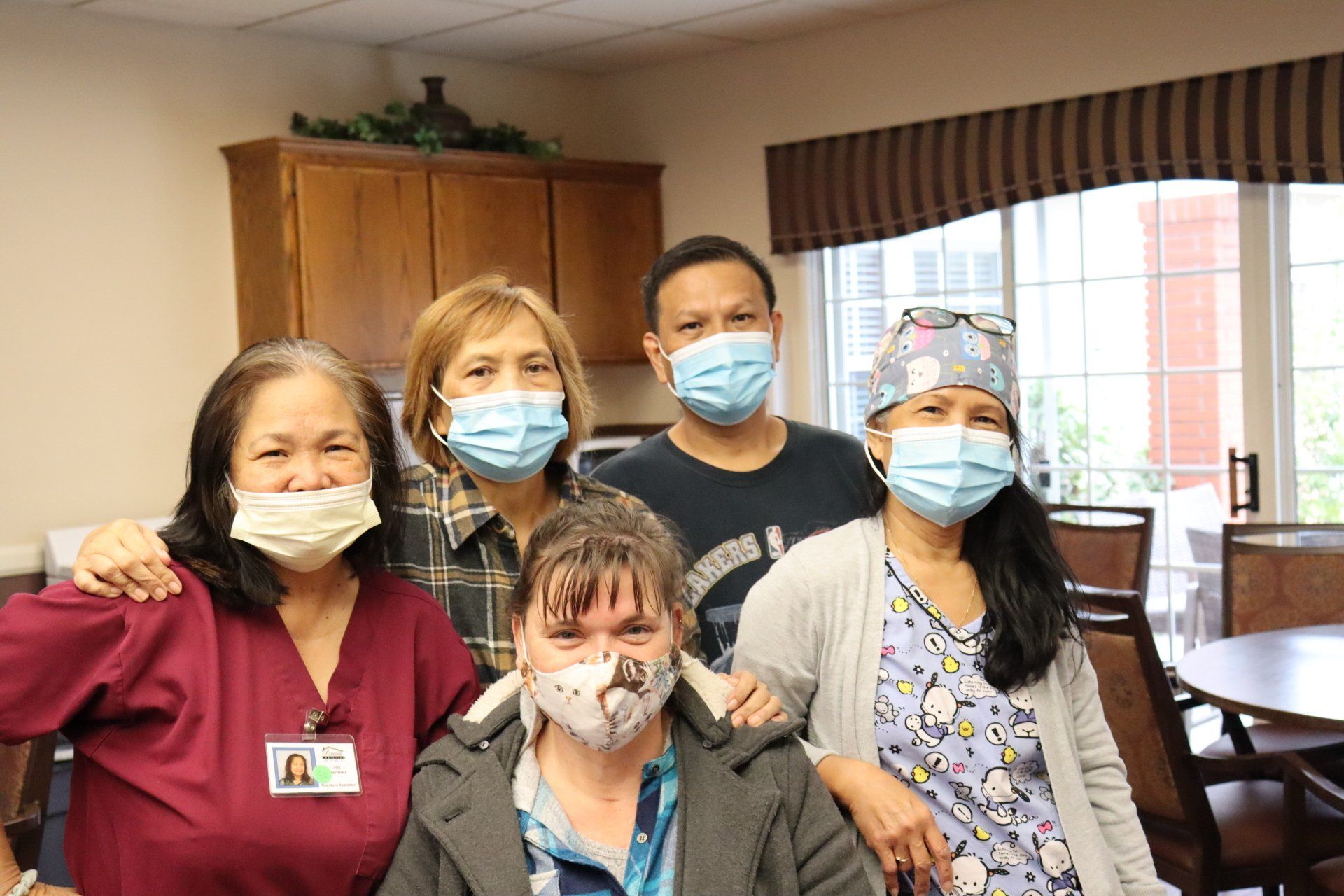 Five masked people pose indoors; two women, one wearing scrubs, and a man in front of a dining area.