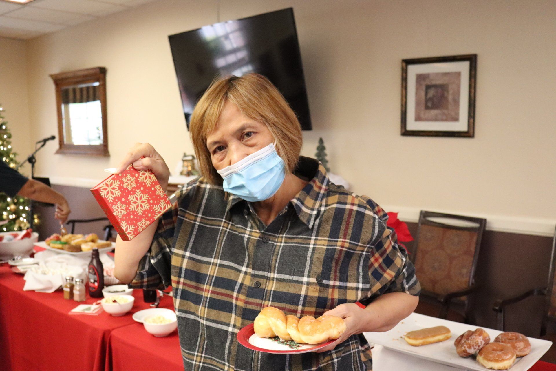 Woman with mask holds plate of donuts, near a Christmas tree and snacks.