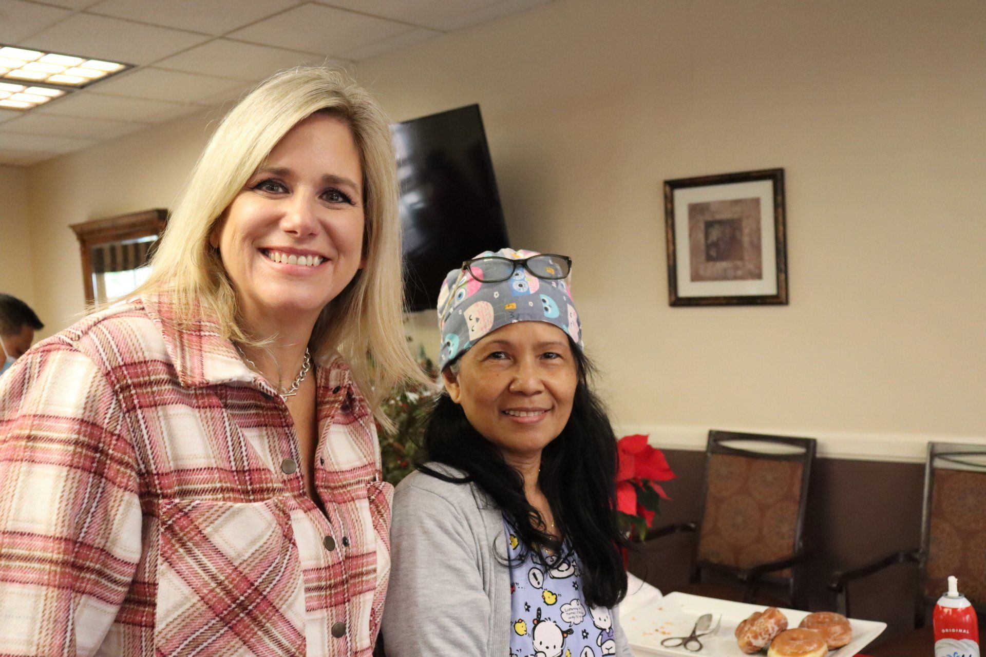 Two women smiling indoors. One wears a plaid shirt, the other a surgical cap.