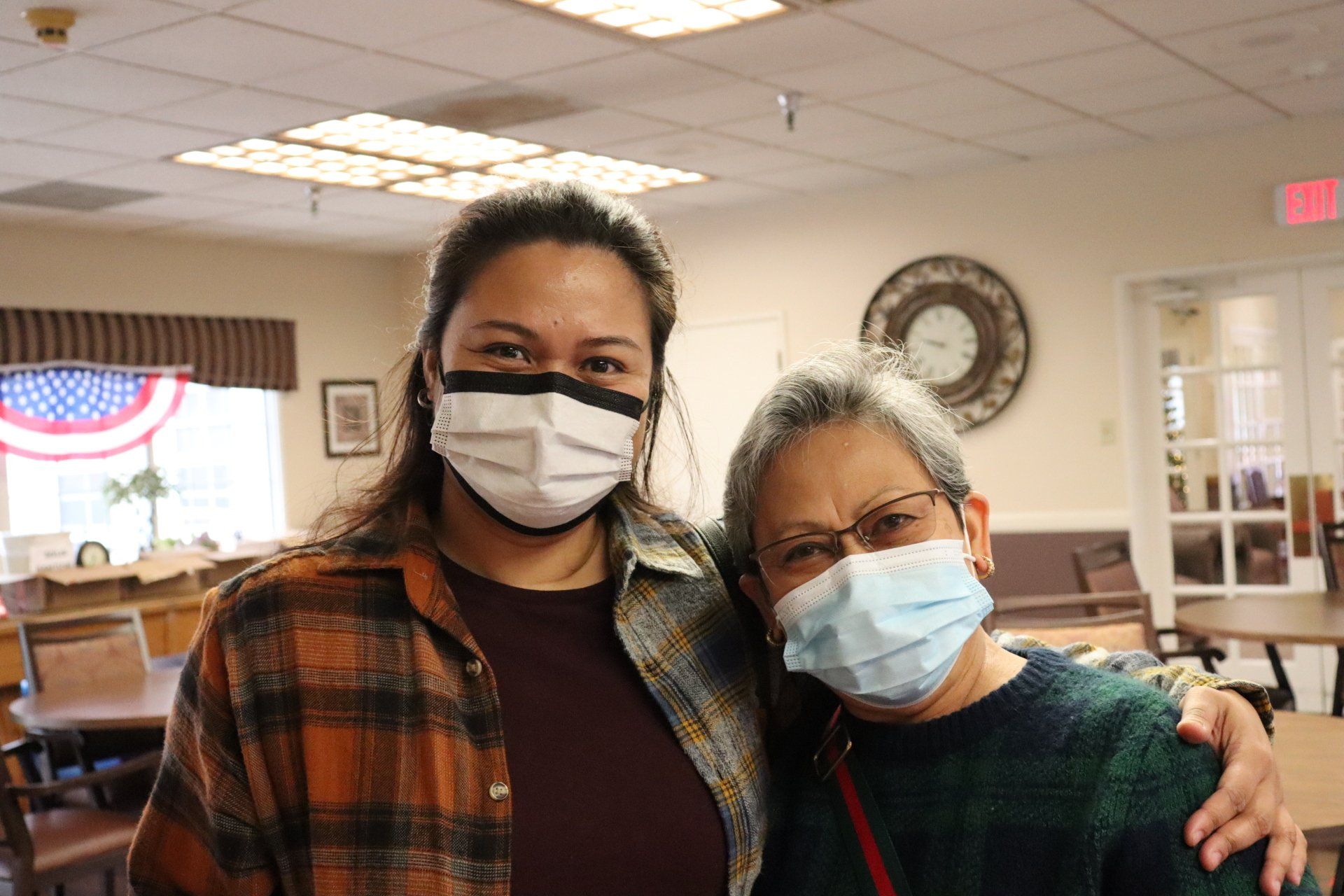 Two women in masks embrace indoors. One wears plaid, the other glasses and a sweater.