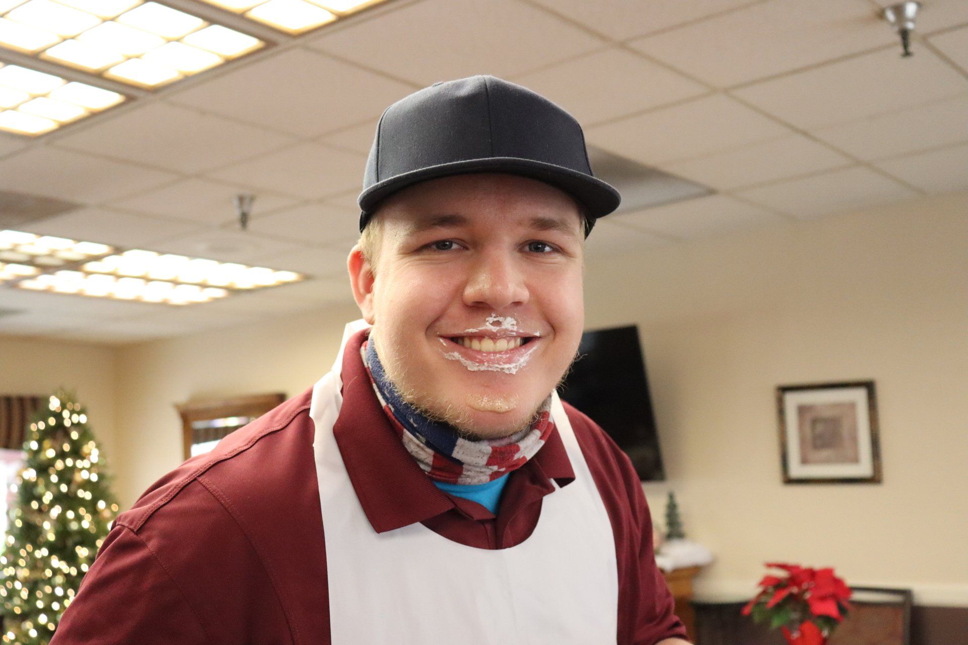 Man with frosting on his face wearing a black cap and apron in a decorated room.