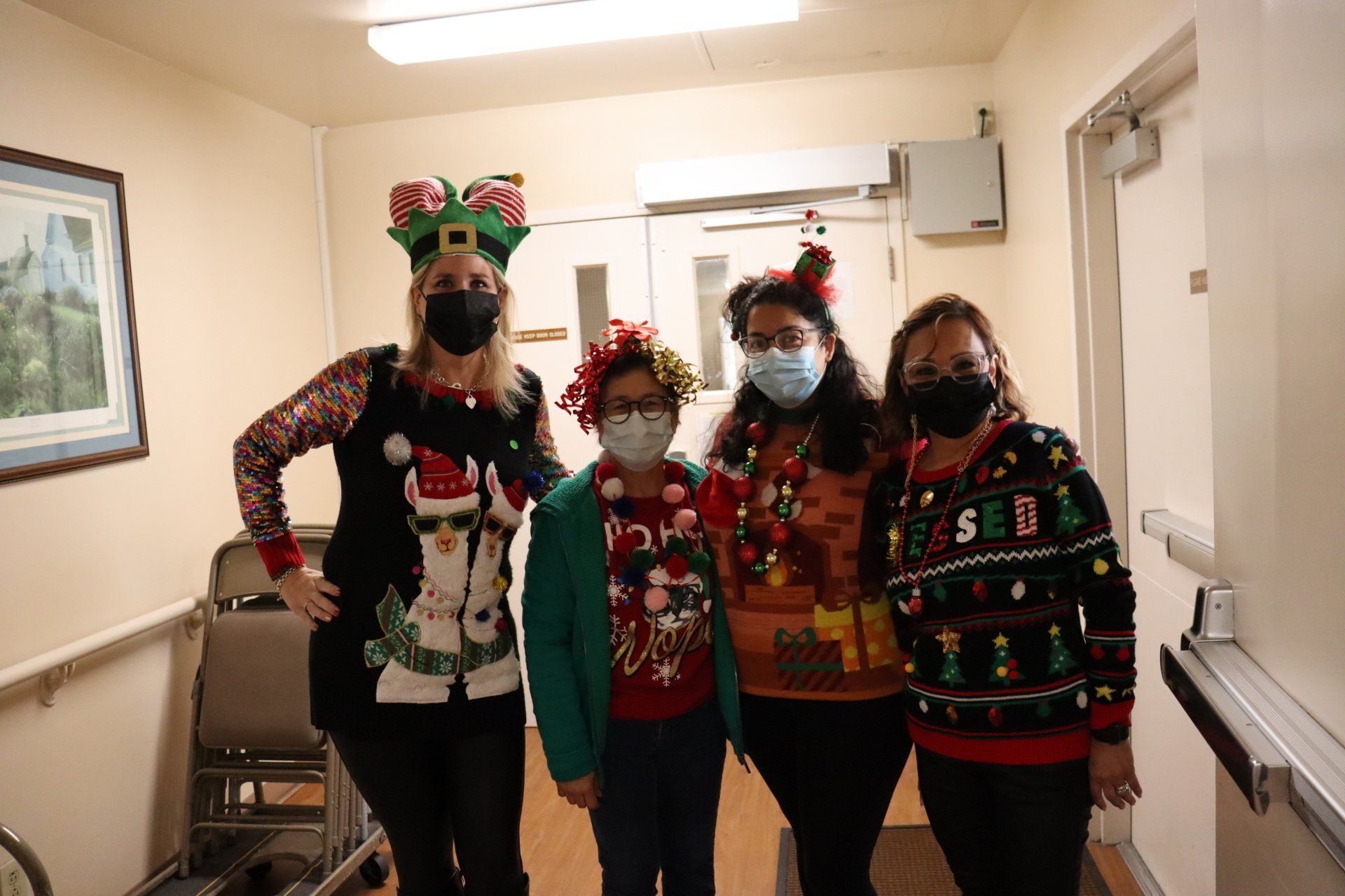 Four people in Christmas sweaters and masks pose in a hallway.