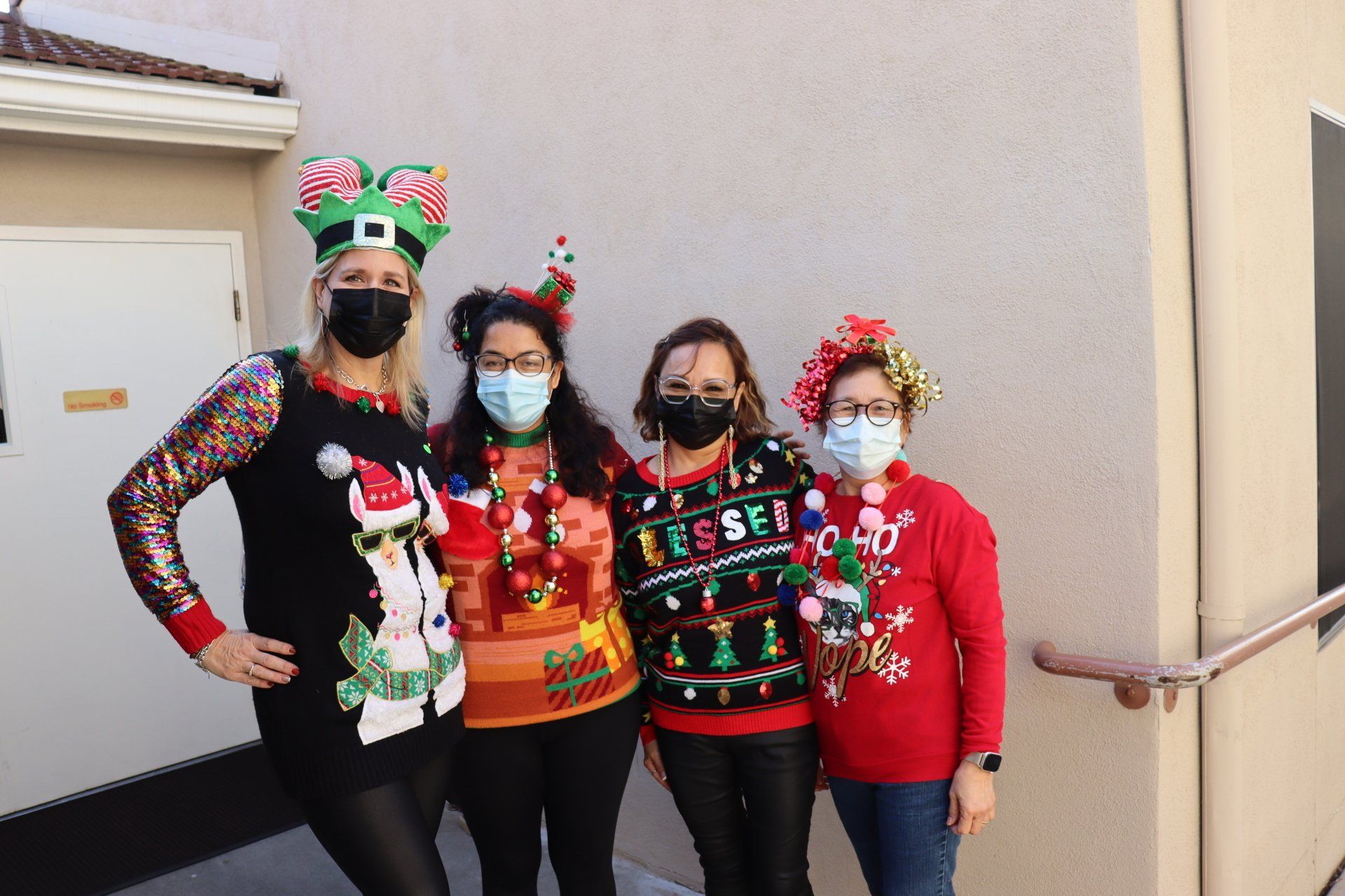 Four people in festive Christmas sweaters and masks posing outdoors.