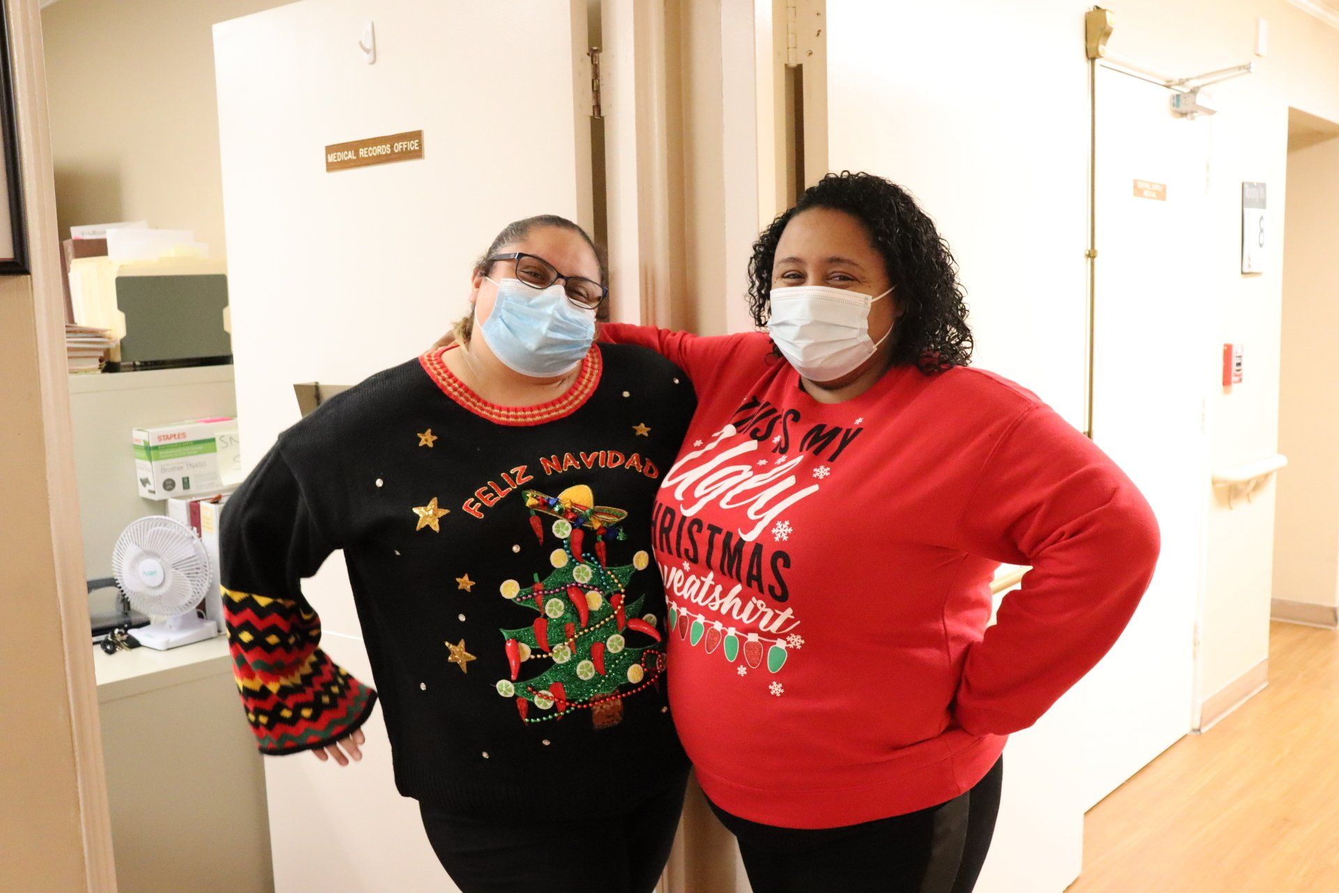 Two women in Christmas sweaters and masks pose in a hallway. One has an arm around the other.