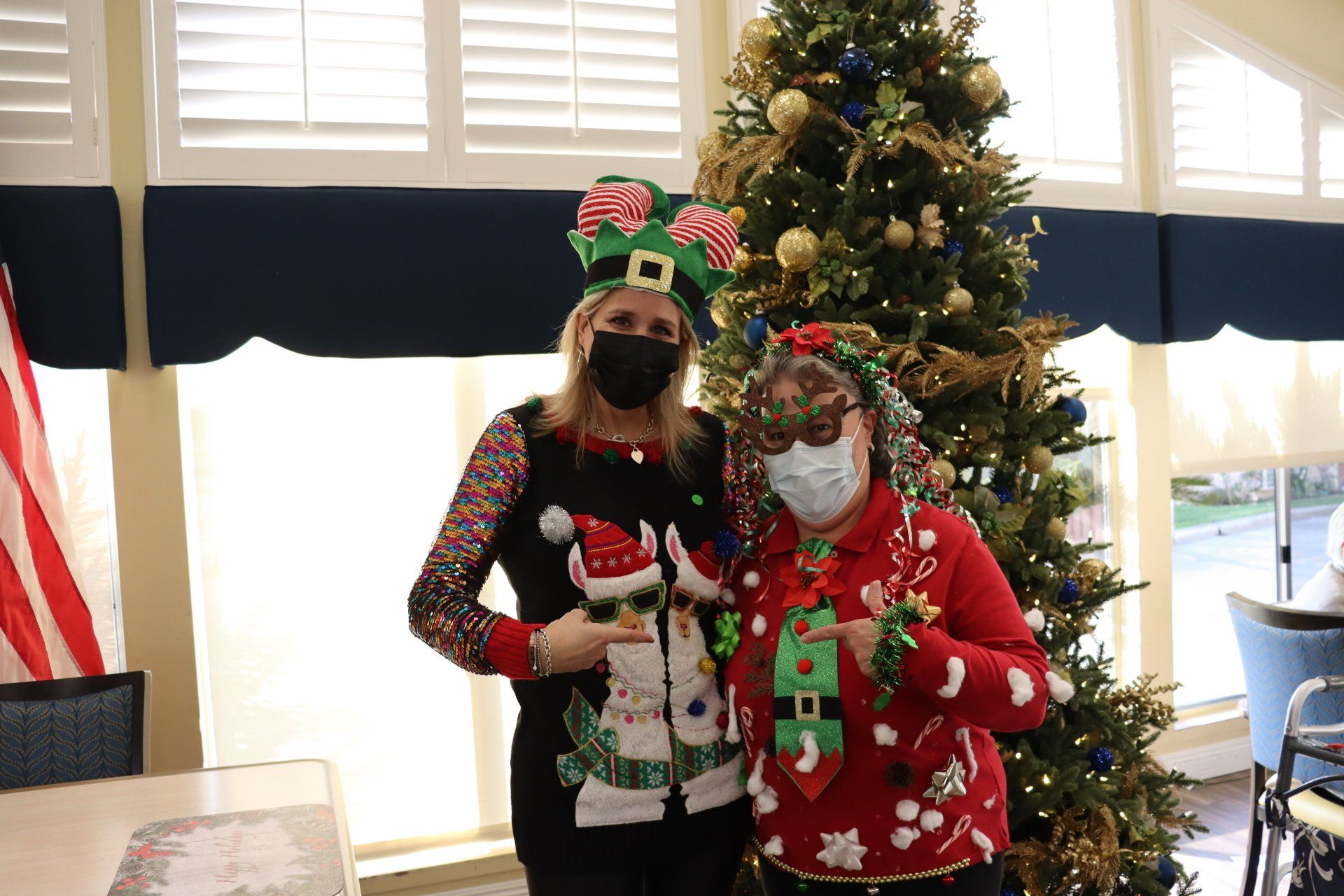 Two people in festive sweaters and masks pose by a Christmas tree indoors.