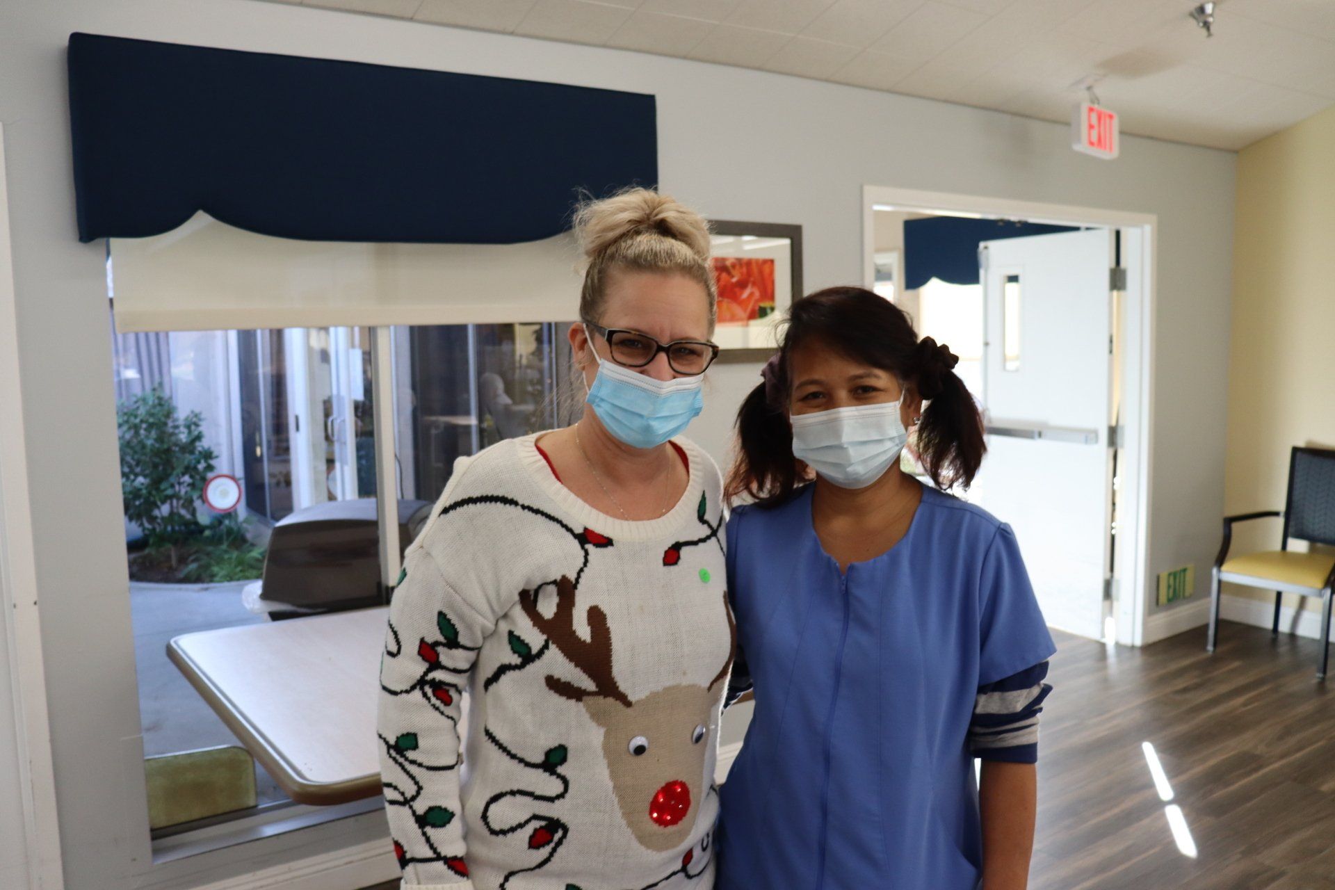 Two women wearing masks, one in a reindeer sweater, the other in blue scrubs, pose indoors near a window.