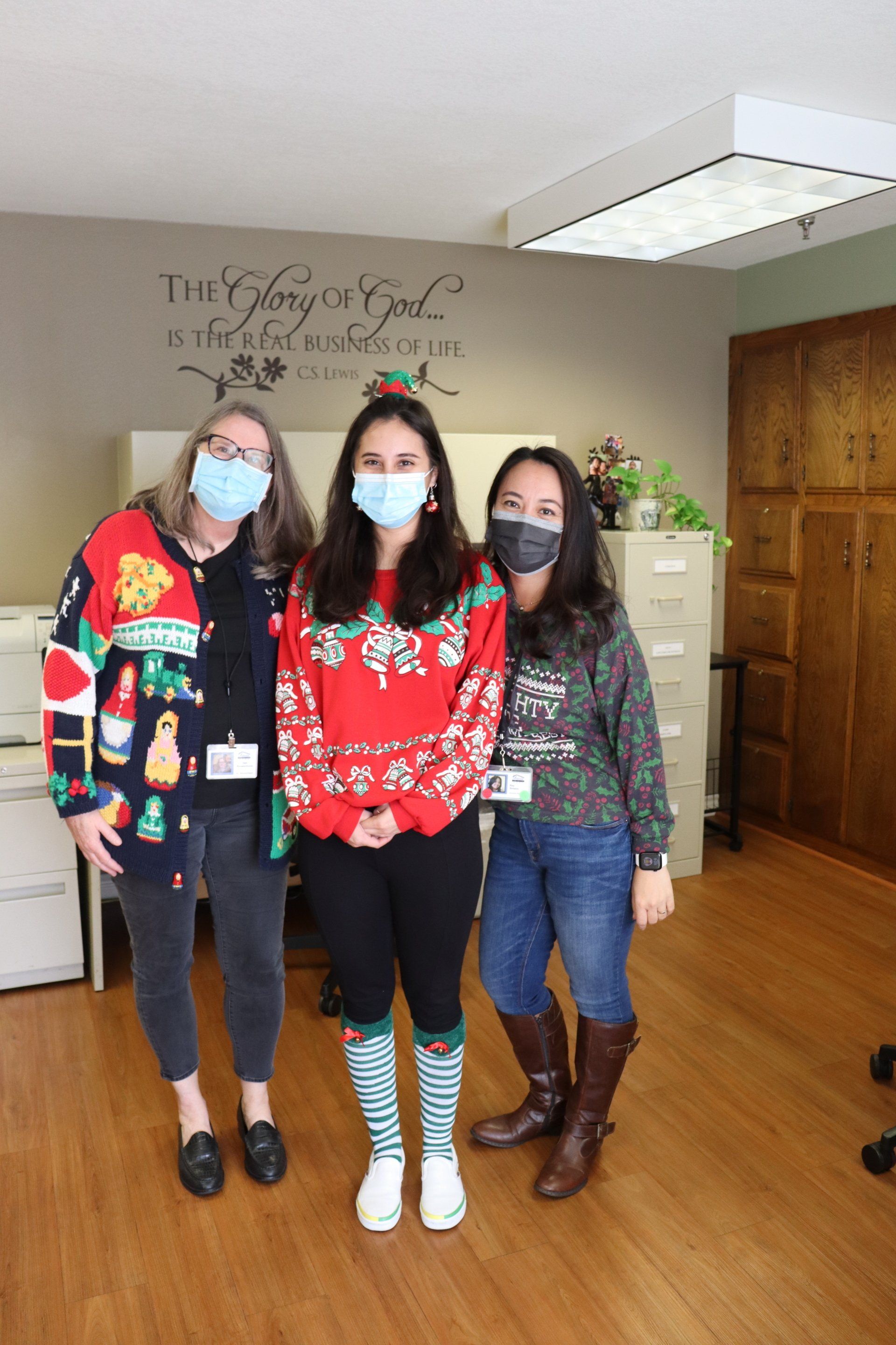 Three people in festive sweaters and masks pose in an office.