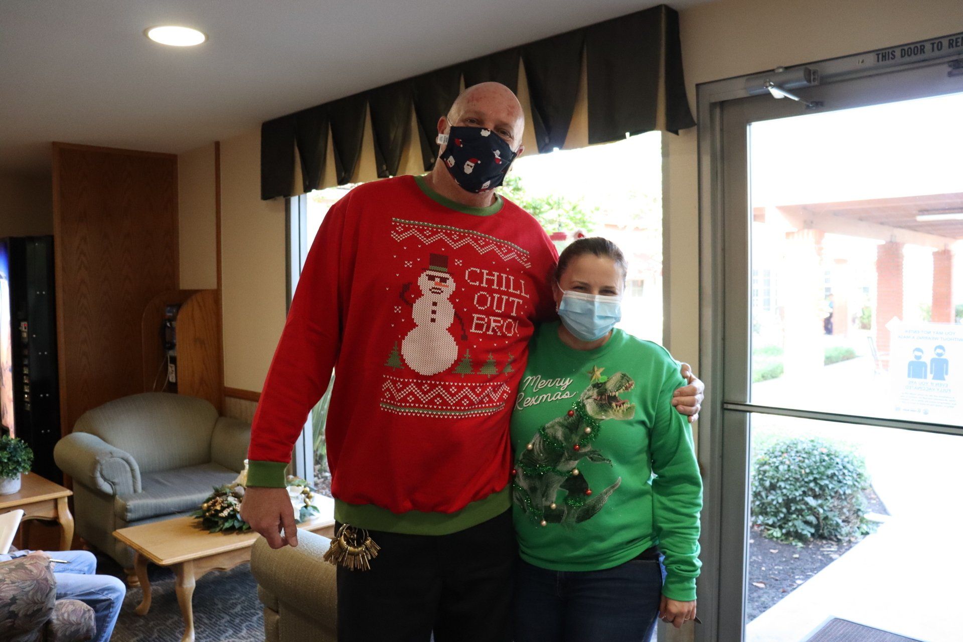 Two people in holiday sweaters and masks pose indoors.