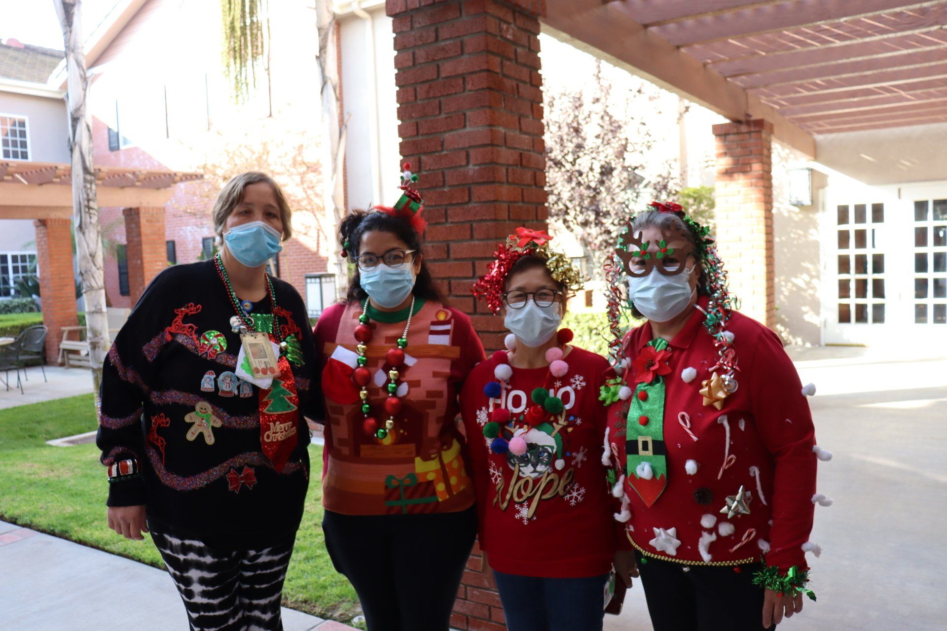 Four people in festive sweaters and masks pose outdoors, smiling, with a brick building in the background.