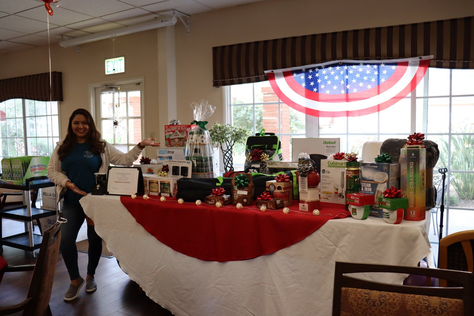 Woman stands beside a table covered in gifts; room with American flag, festive decor.