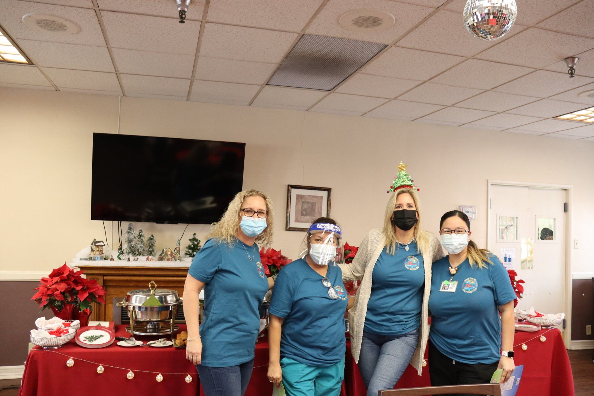 Four healthcare workers in teal shirts and masks pose at a Christmas table in a decorated room.