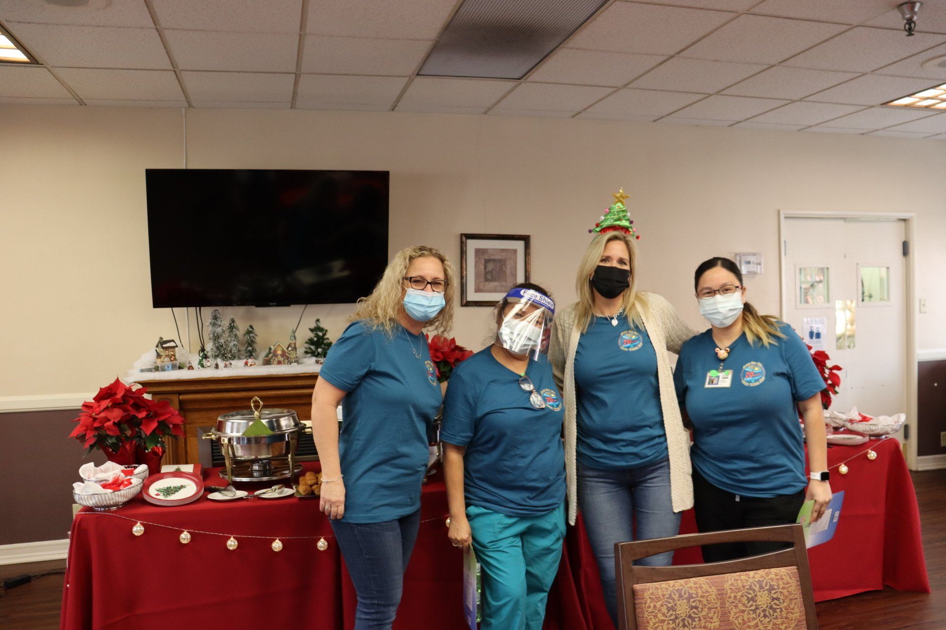 Four masked people in teal shirts and jeans stand near a table with Christmas decorations.