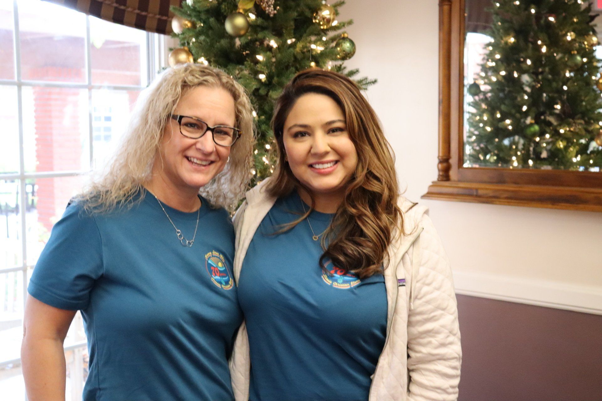 Two women smiling, wearing teal shirts, in front of a Christmas tree.