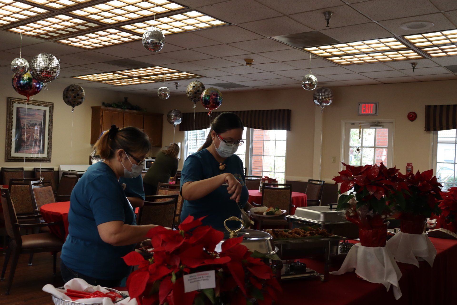 Two masked people prepare food at a holiday buffet, poinsettias and disco balls decorate the room.