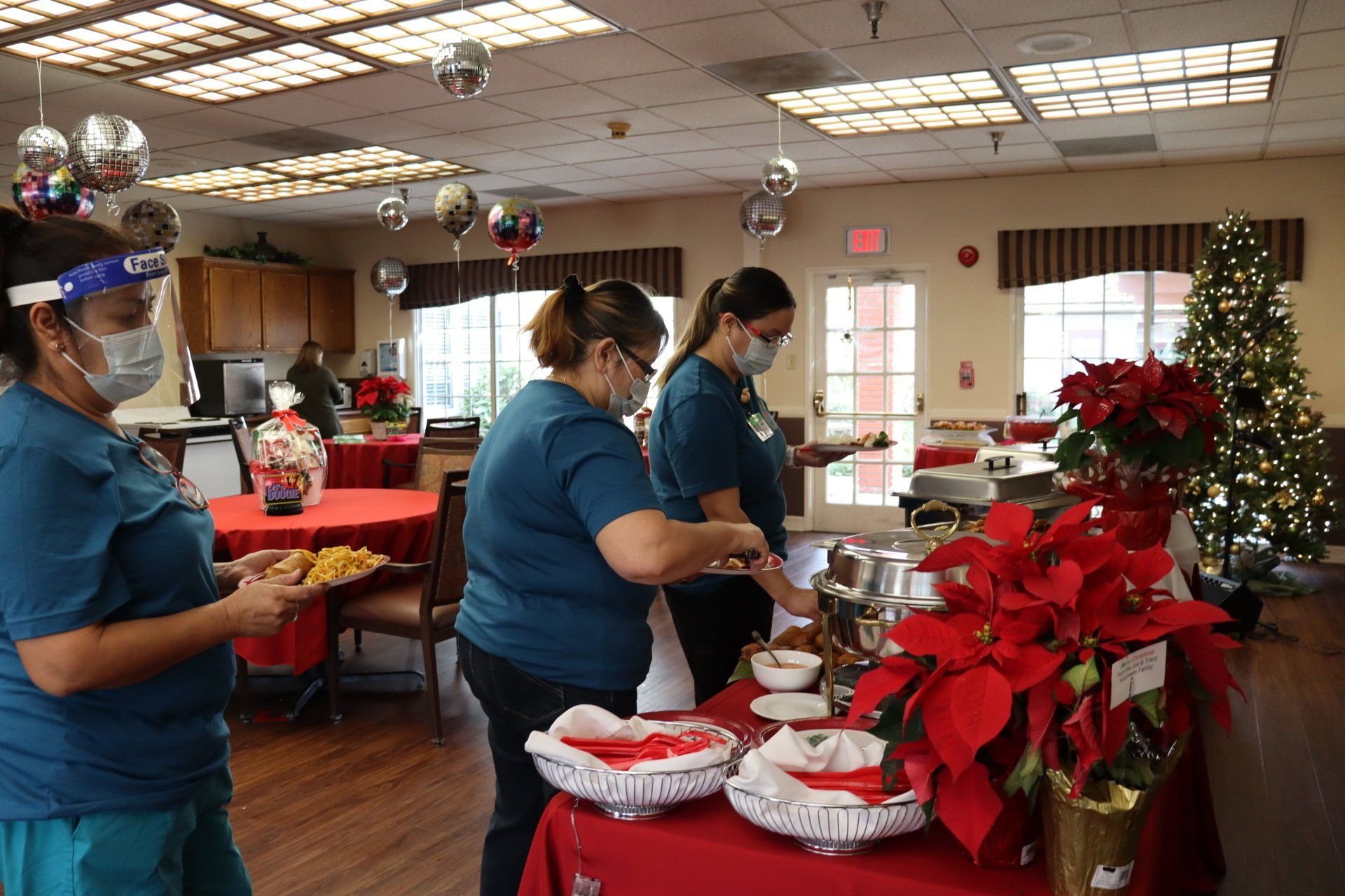Three masked healthcare workers serving food at a holiday buffet in a decorated dining room.