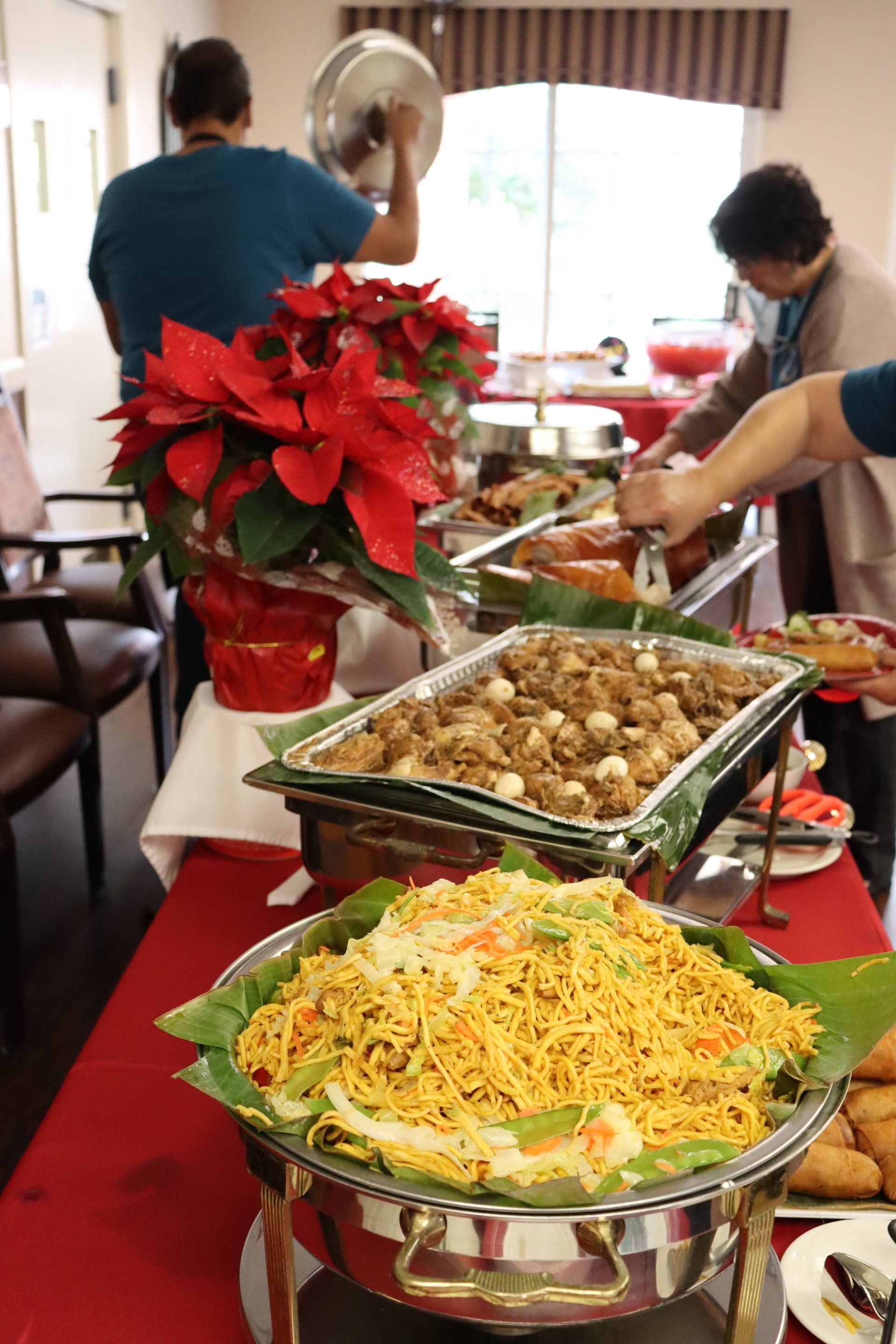Buffet with various dishes, red tablecloth, people serving food, poinsettia arrangement.
