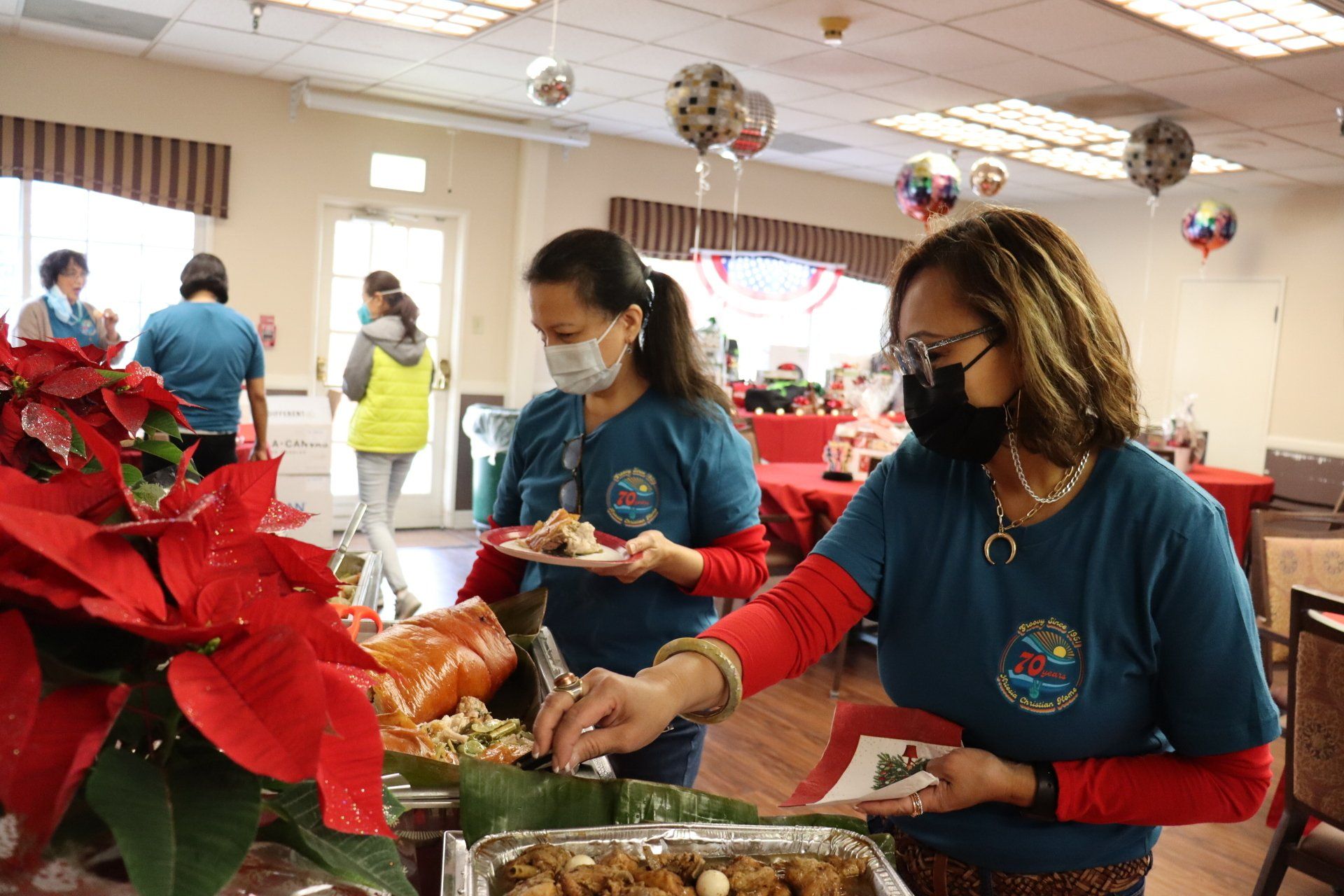 Two women serve food at a buffet. One wears a face mask. Festive decorations are in the background.