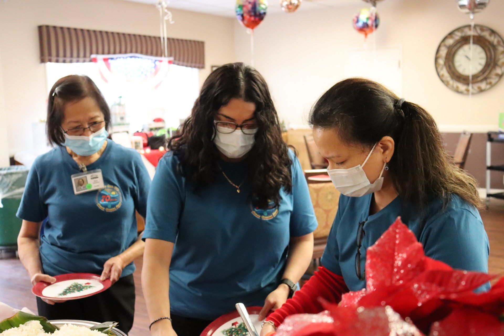 Three women in blue shirts and masks serving food at an event; red poinsettia, balloons, clock visible.