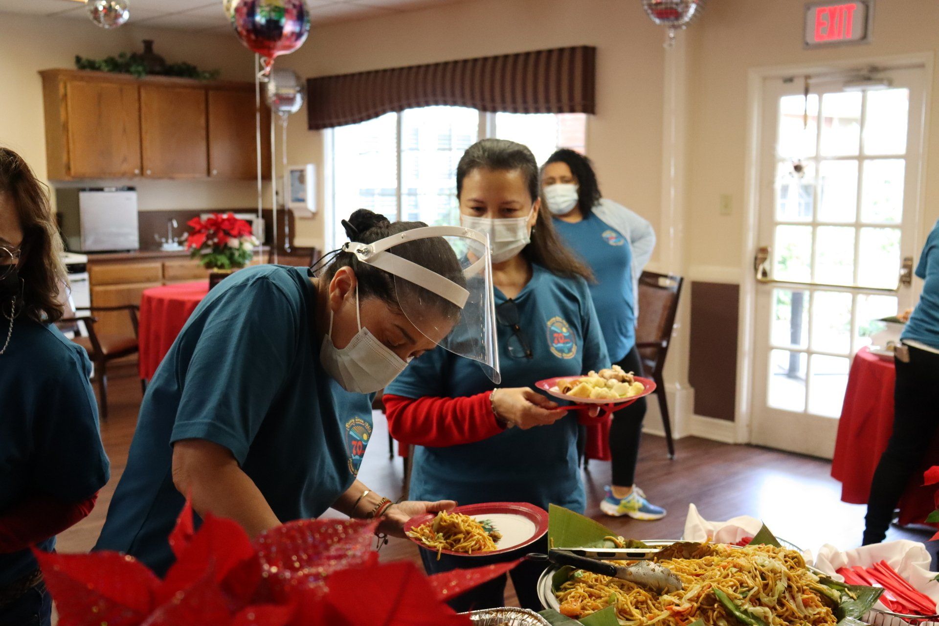 Staff in blue scrubs serve food at a catered event, with masks, shields, and festive decorations.