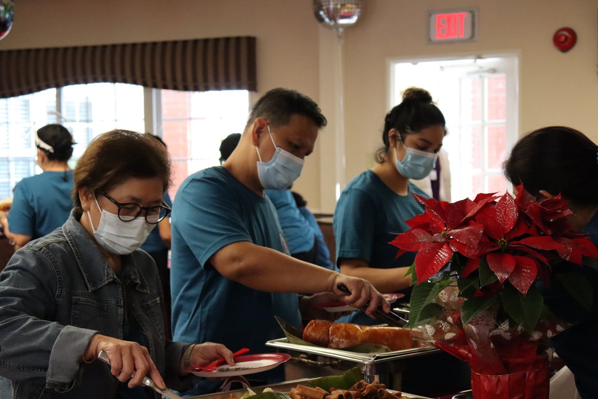 People wearing masks at a buffet serving food, indoors with red poinsettia.