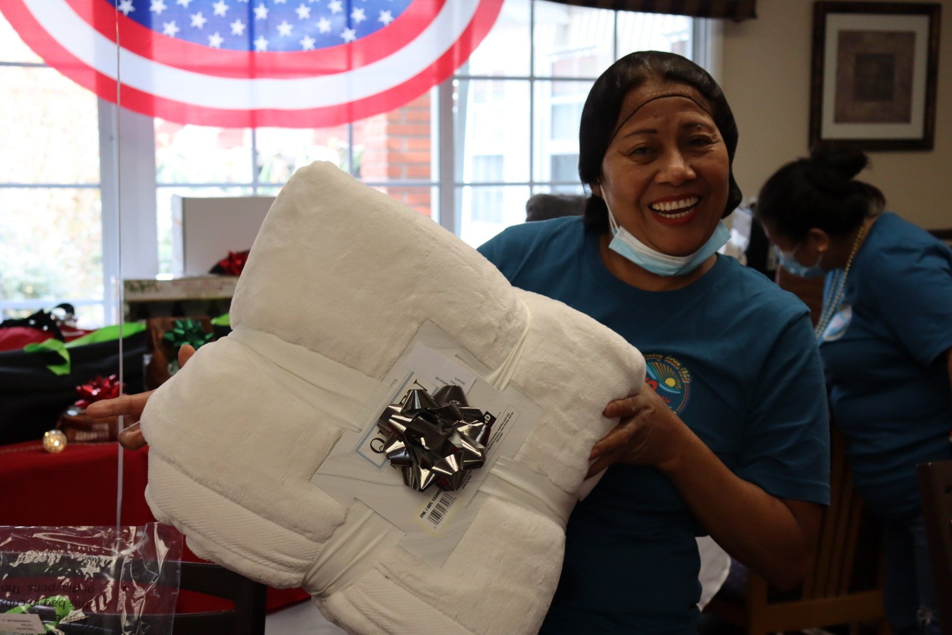 Woman holding a white, gift-wrapped blanket; smiles in a room with an American flag and another person.