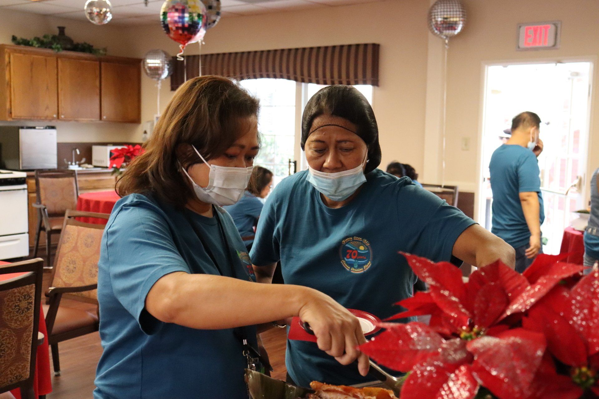 Two women in masks serving food at an event, red poinsettias in foreground.