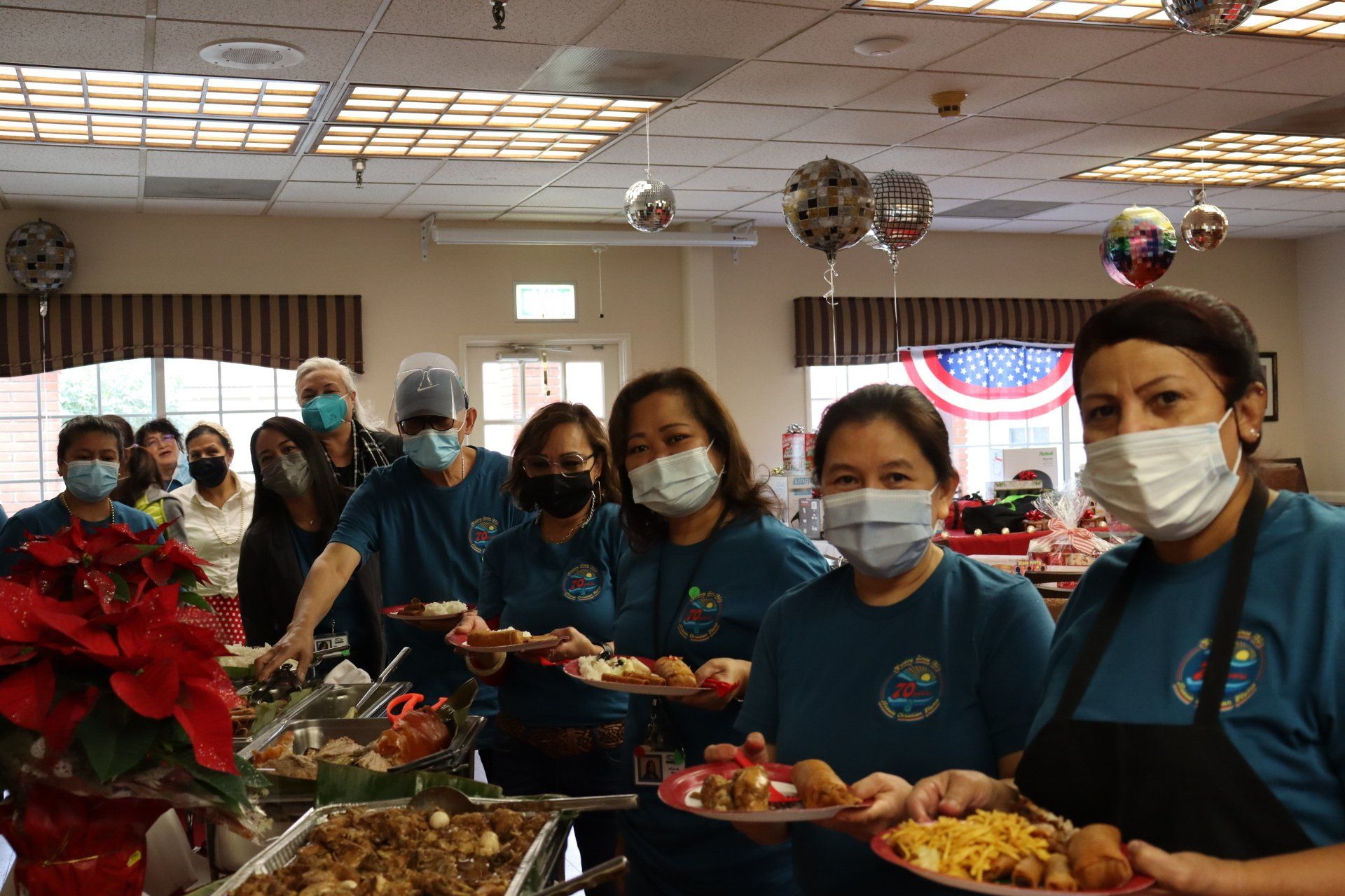 Group of people serving food at an event.  Most are wearing masks and matching teal shirts. Festive decorations visible.