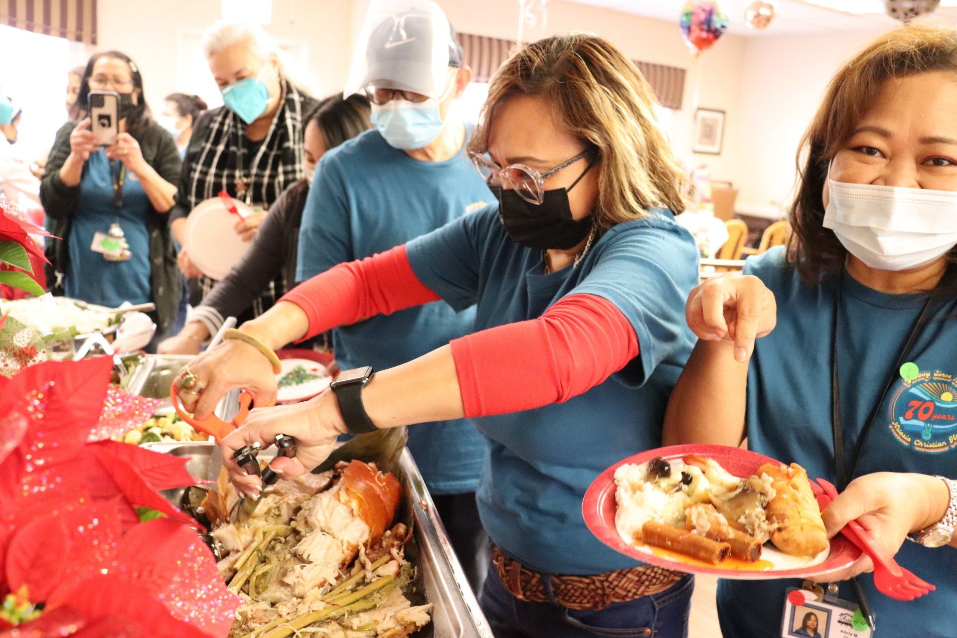 People in masks serving food at a buffet, festive decorations, some holding plates, indoor setting.