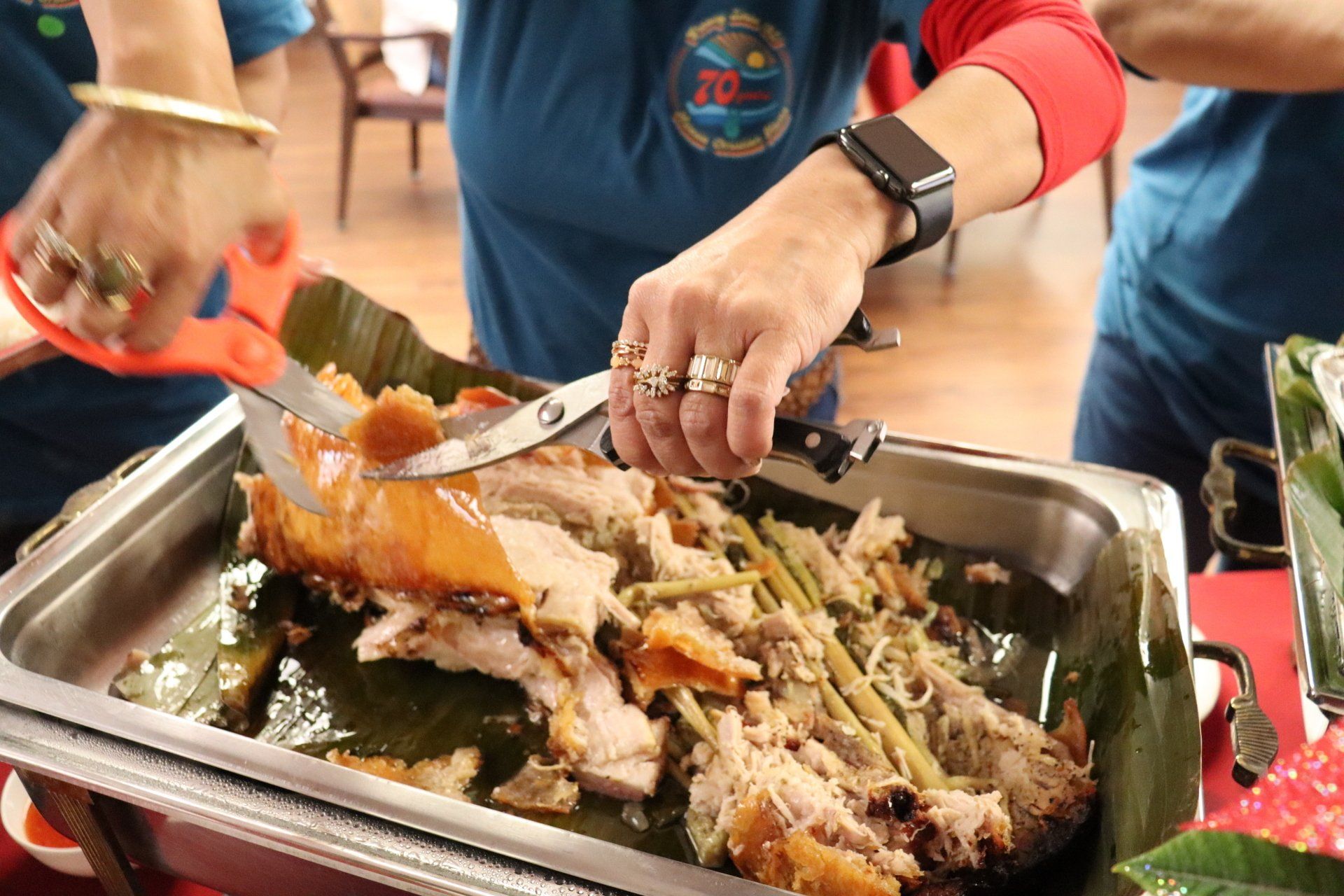 Person cutting roasted pork with scissors in a silver pan lined with banana leaves.