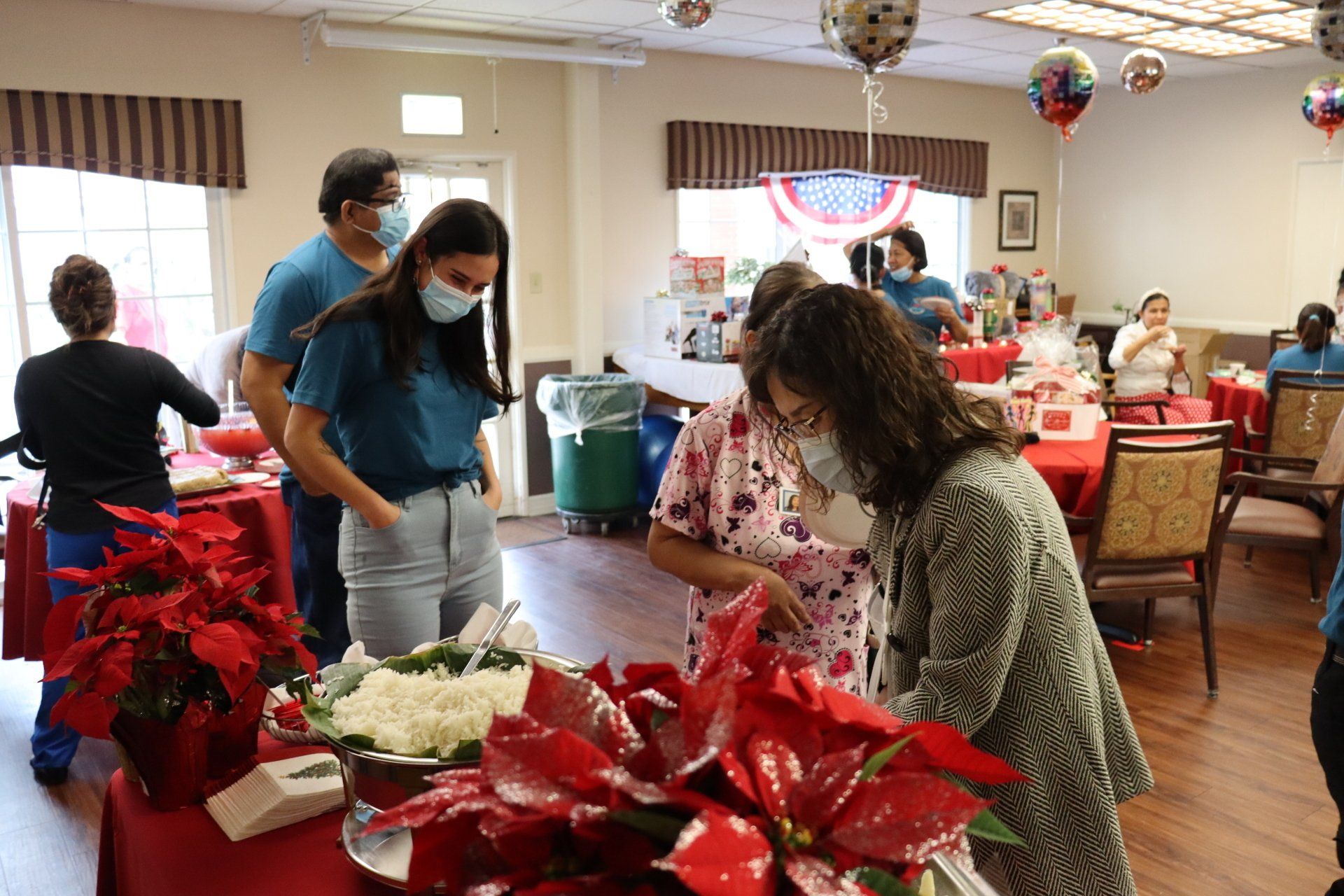 People in masks at a decorated event with food, red poinsettias, and patriotic decorations.