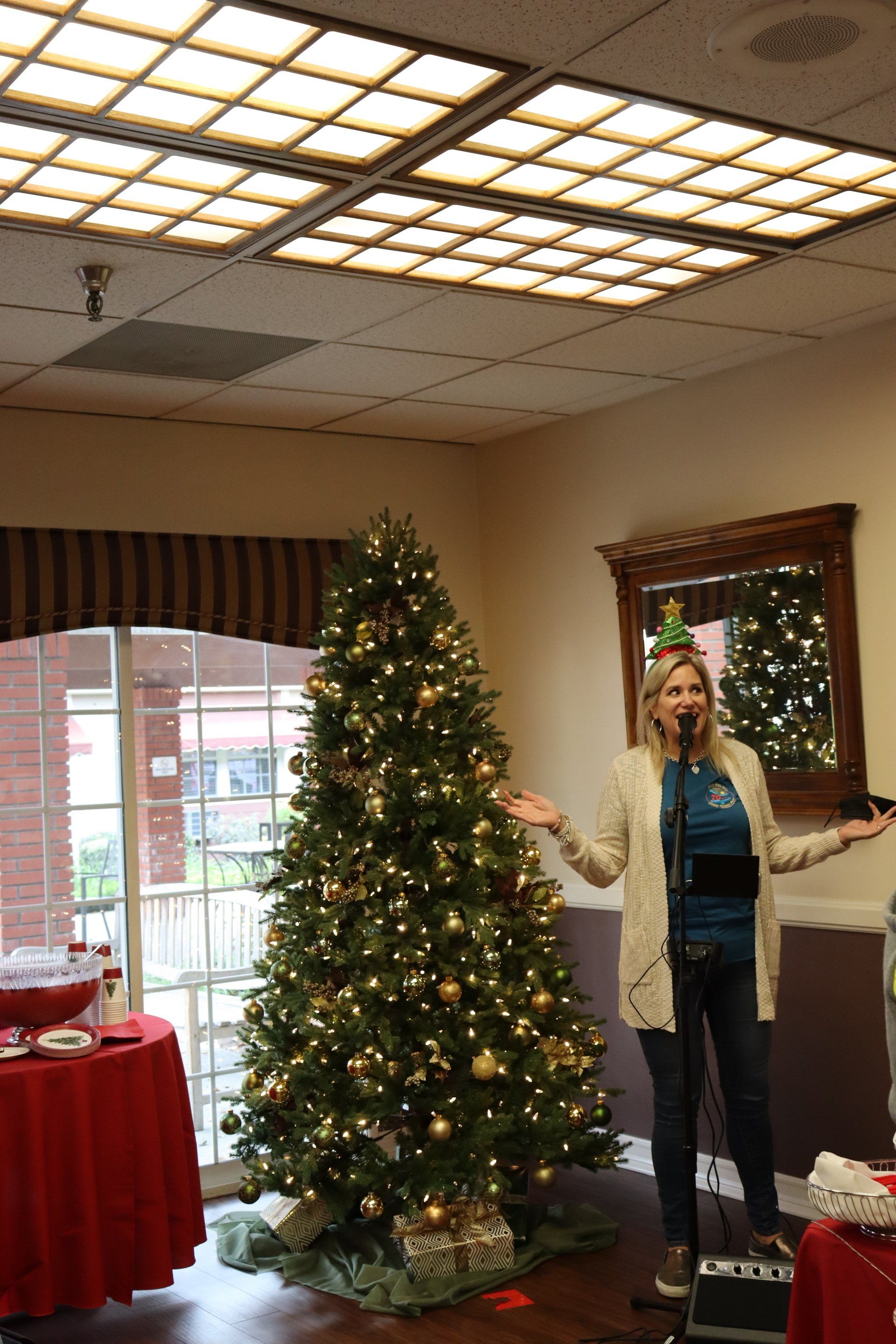 Woman singing, Christmas tree, microphone, party. Beige walls, glass door, and red table.