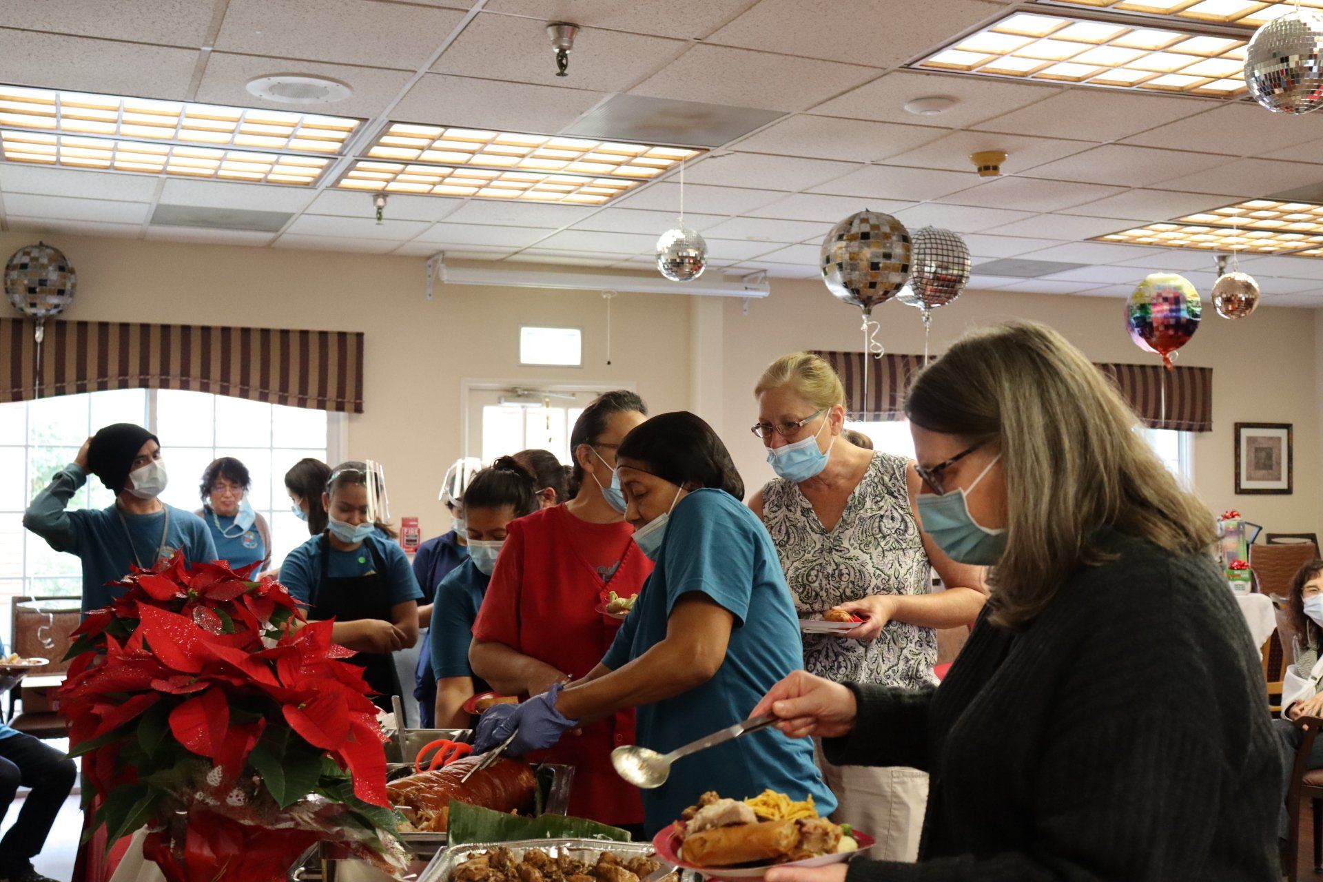 People serving food at a buffet; red poinsettia centerpiece; masks; balloons.