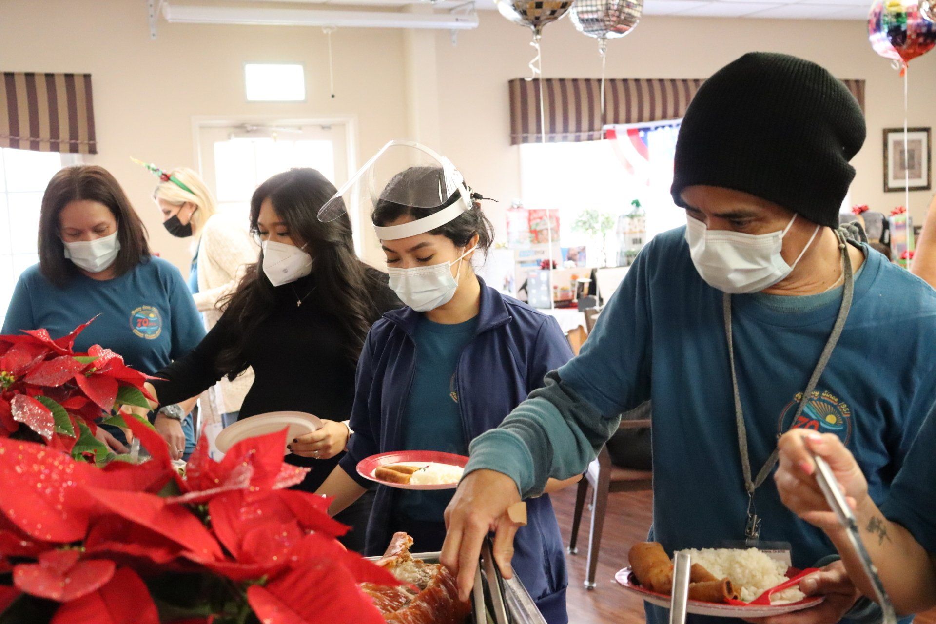 People in masks serving food at a buffet table decorated with red poinsettias.