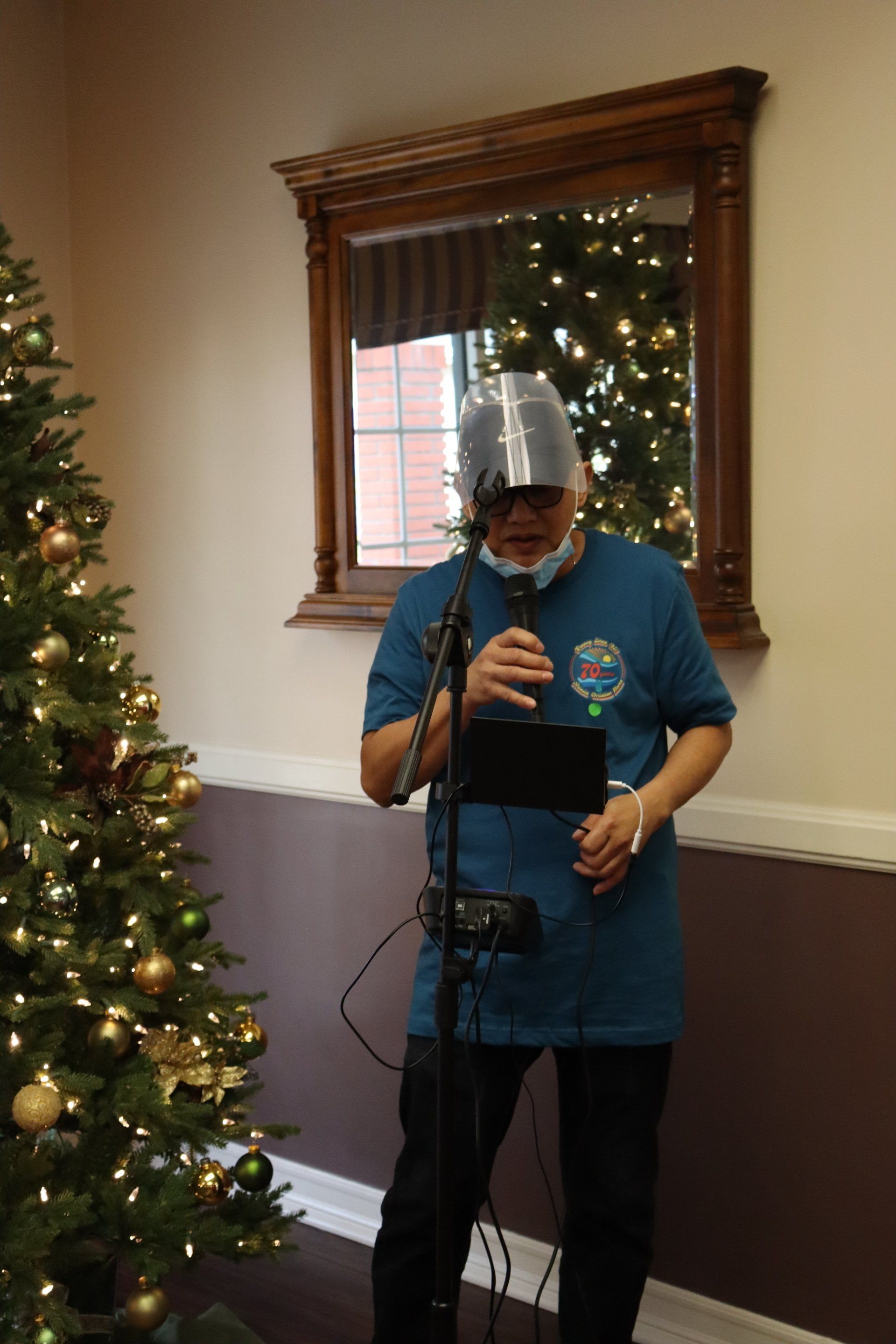 Man in a teal shirt with a face shield, speaking into a microphone near a Christmas tree and mirror.