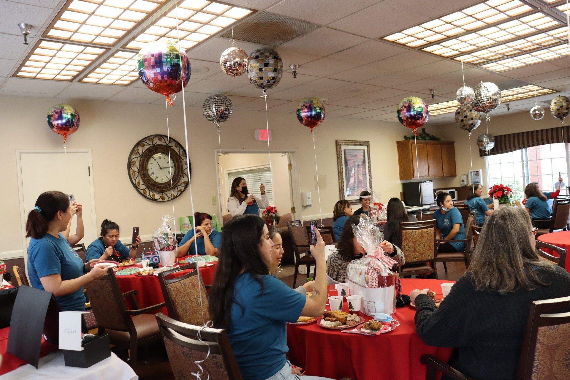 Party in a room with red tablecloths, balloons, and people. Some are taking photos.
